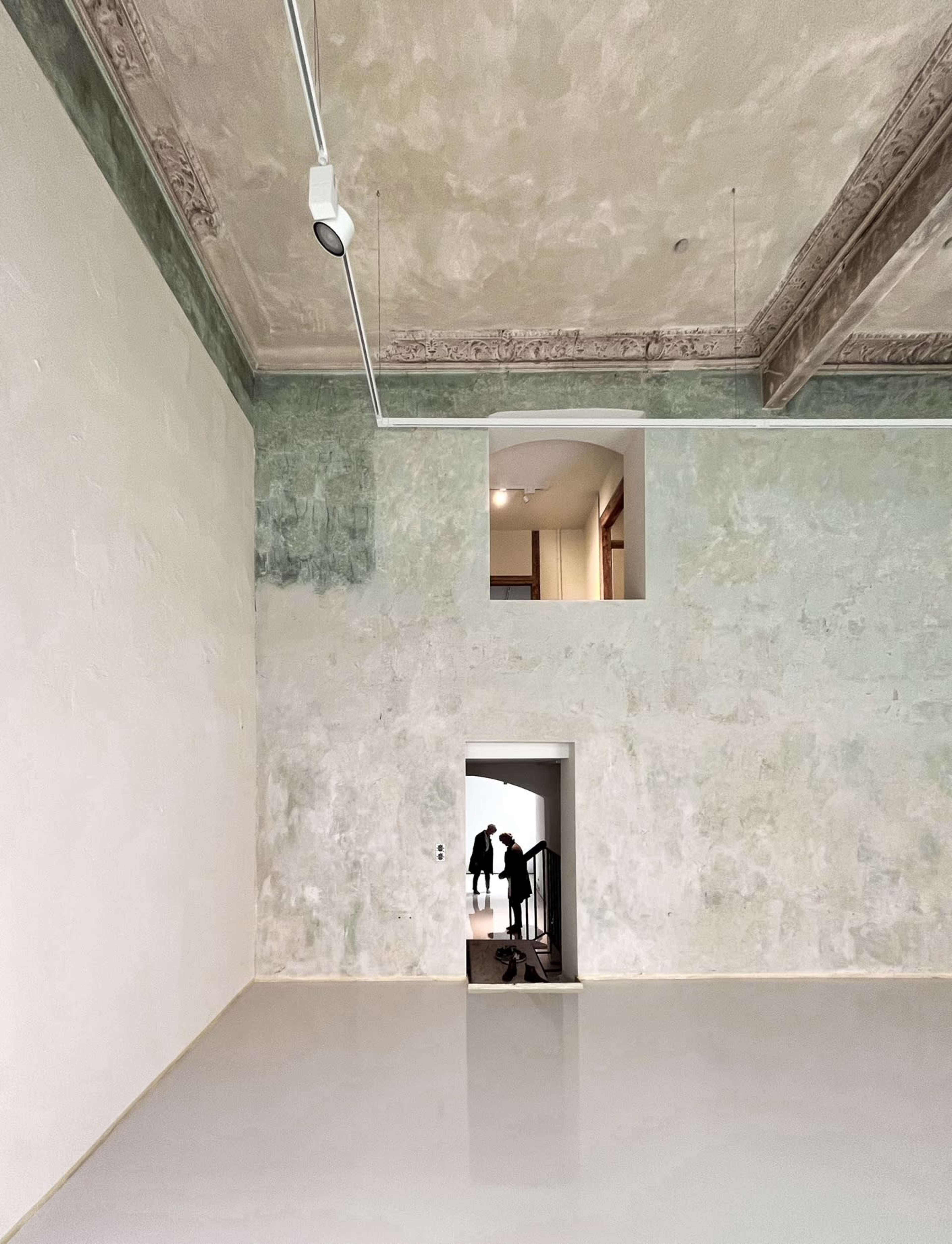 The image depicts a spacious interior with a high ceiling, featuring a dilapidated wall, an arched window, and a doorway leading to a staircase.