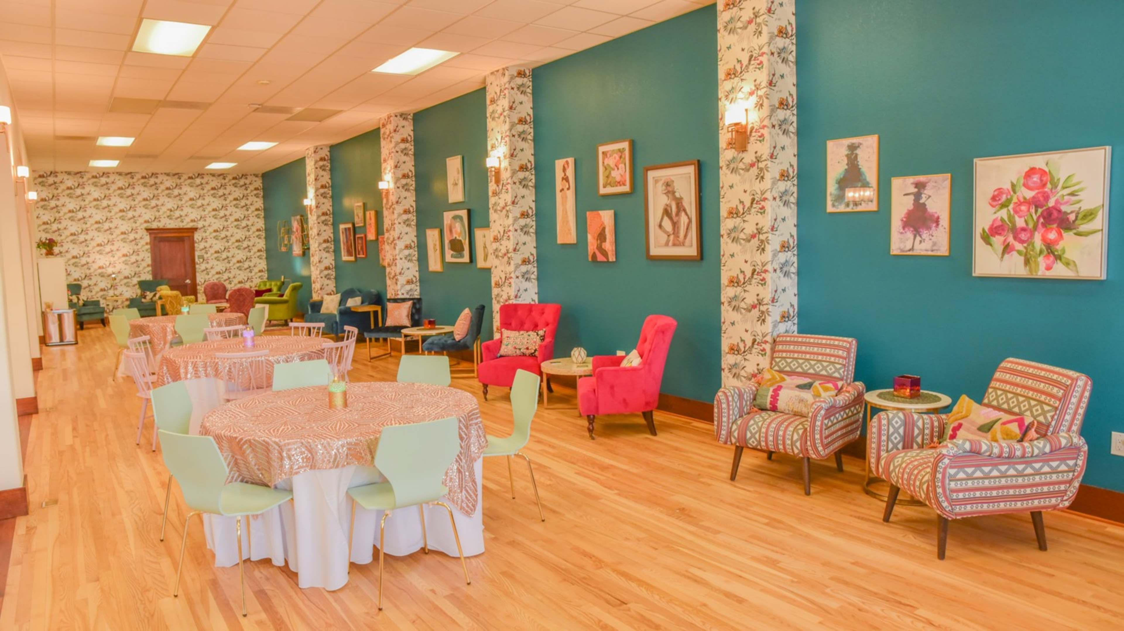 A brightly lit social space featuring tables with floral tablecloths, colorful chairs, and walls adorned with floral wallpaper and framed artwork.