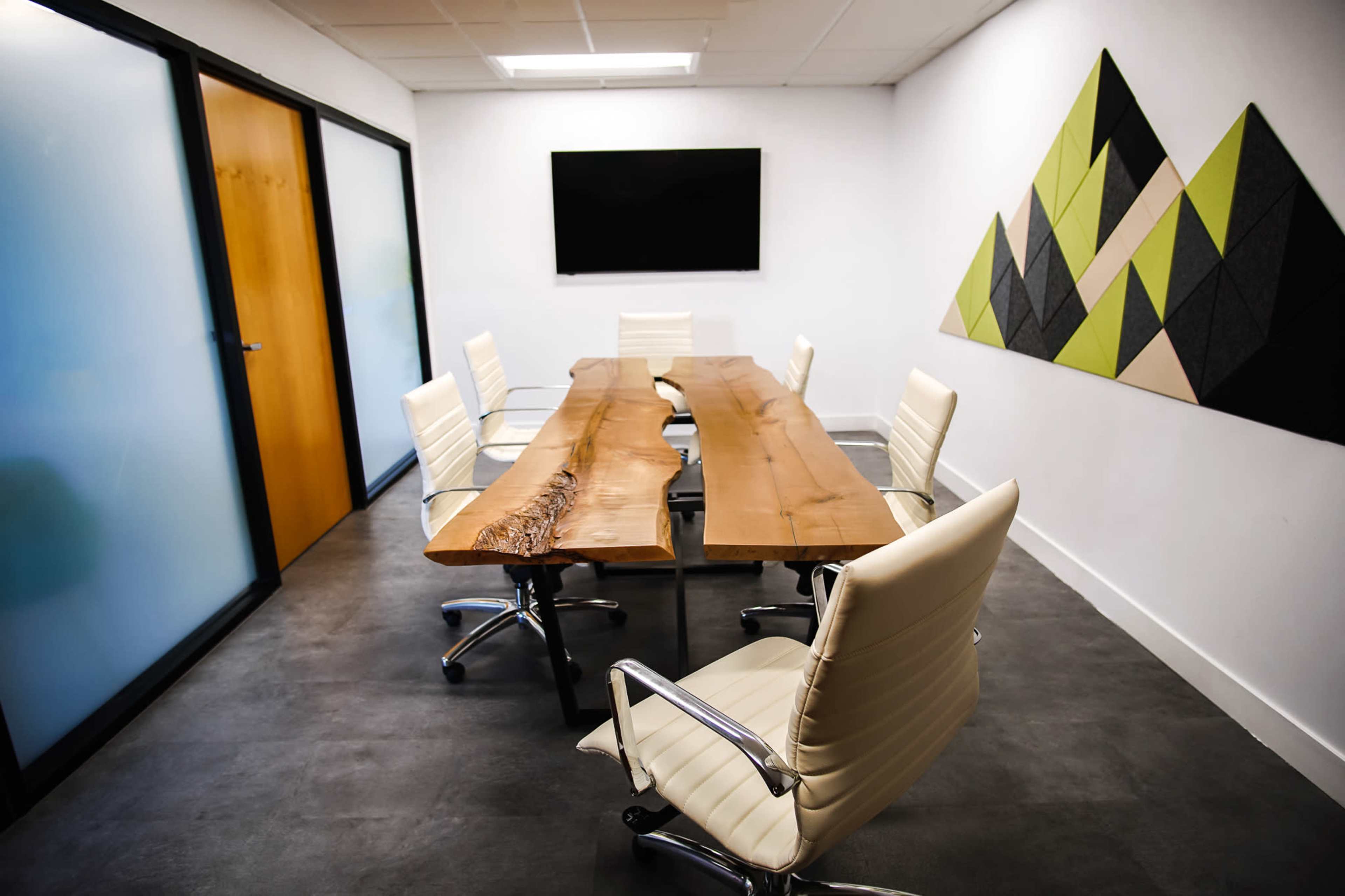A modern conference room features a wooden table surrounded by white chairs, with a large screen mounted on the wall and a geometric design in green and gray.