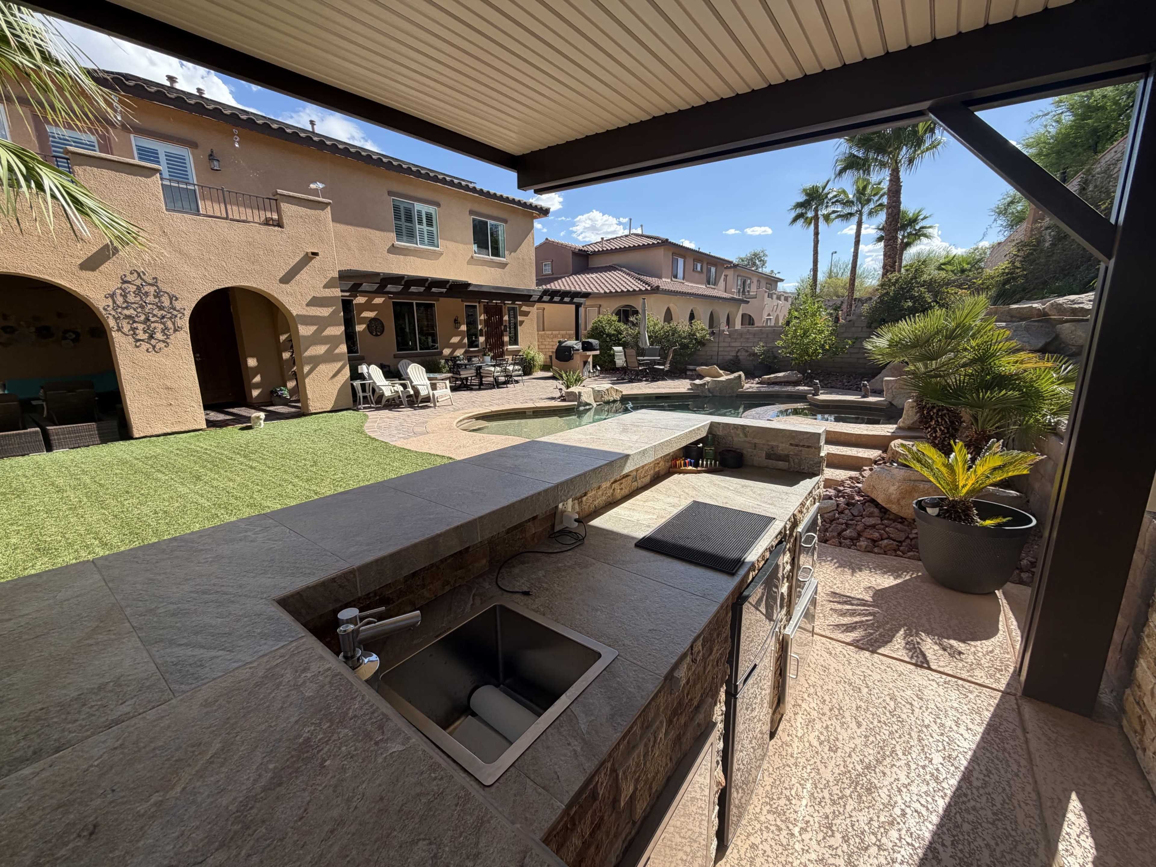 The image shows a spacious backyard with an outdoor kitchen featuring a sink, a swimming pool, and various seating areas surrounded by landscaping and palm trees.
