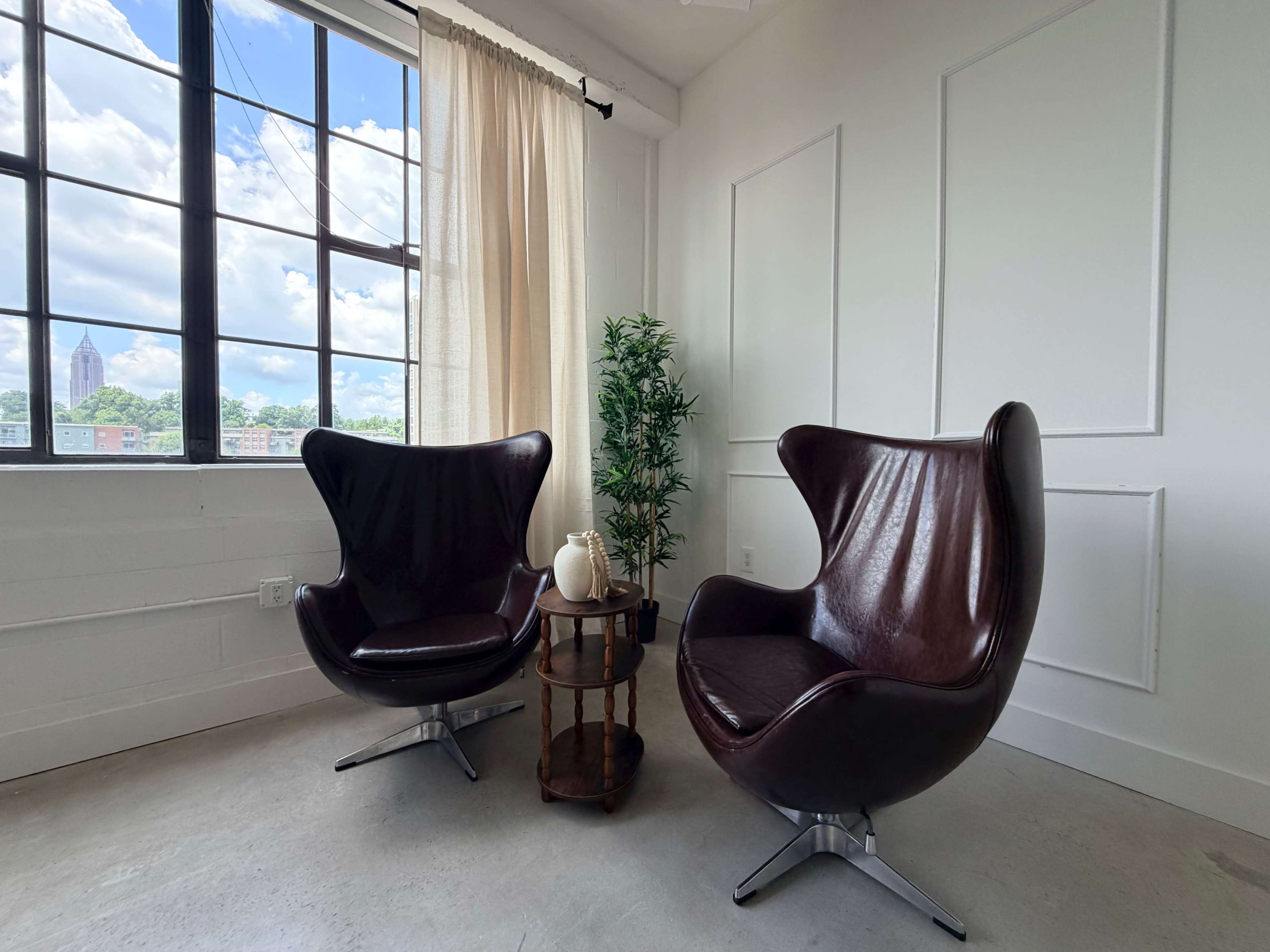 Two modern leather chairs are positioned near a large window with a cityscape view, accompanied by a small wooden table and a potted plant.