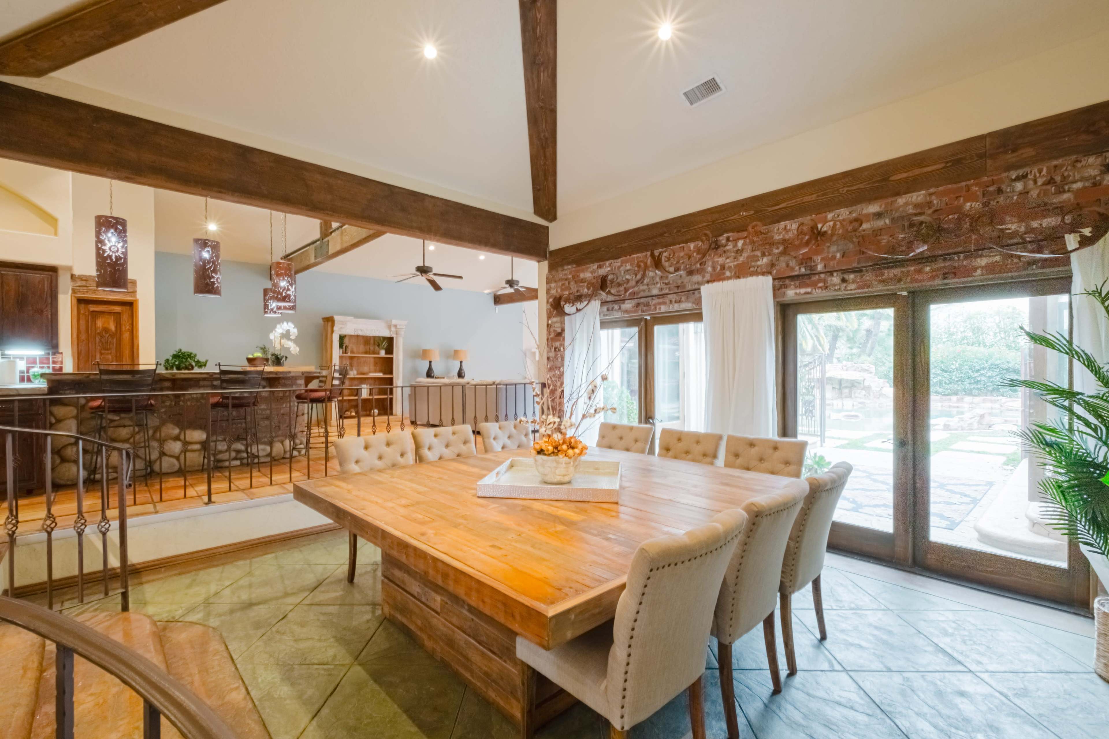A spacious dining area features a large wooden table surrounded by upholstered chairs, with exposed wooden beams and a view of an outdoor patio through glass doors.