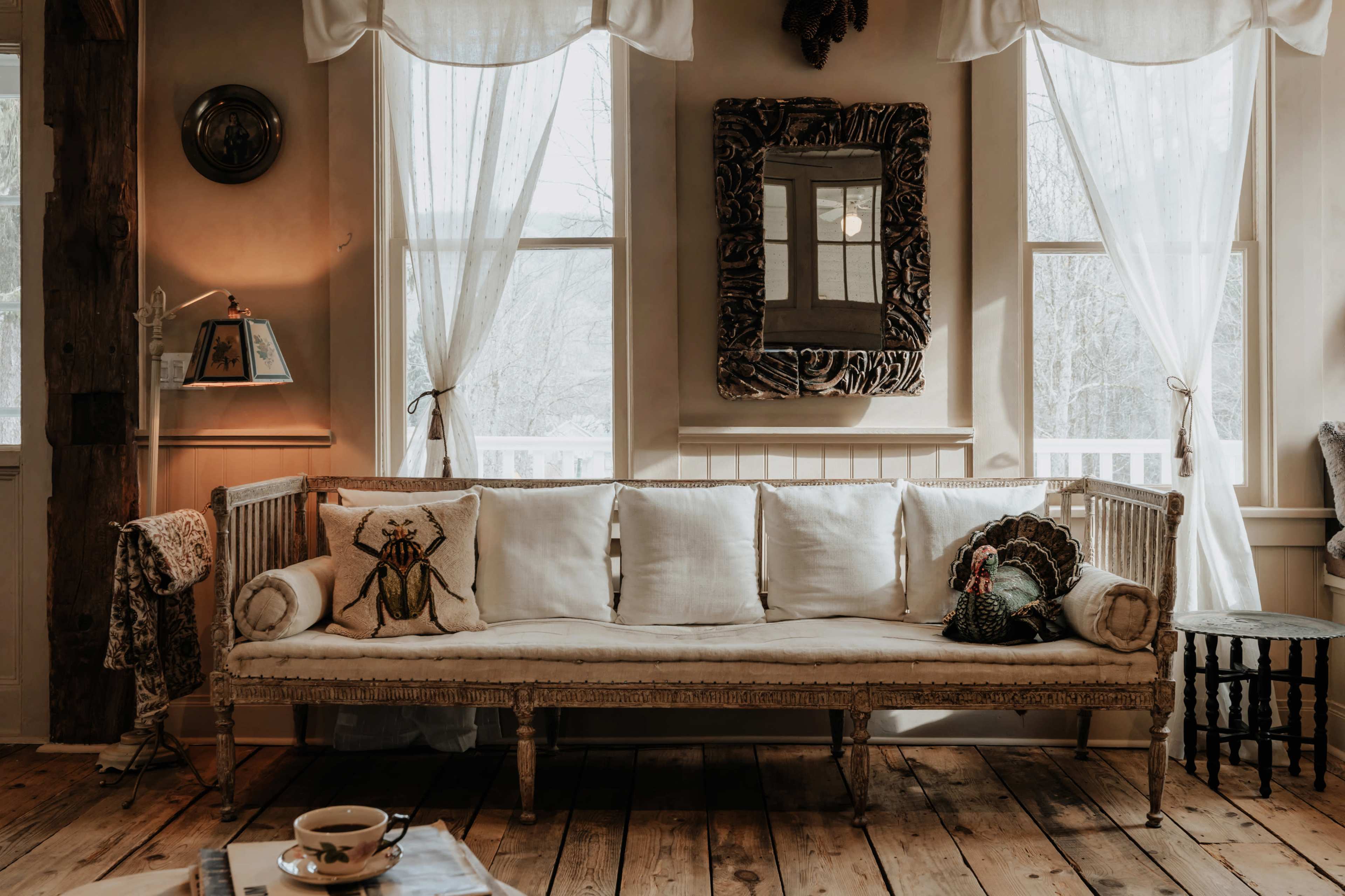 The image depicts a cozy living space featuring a light-colored sofa with multiple cushions, large windows dressed in sheer curtains, and rustic wooden flooring.