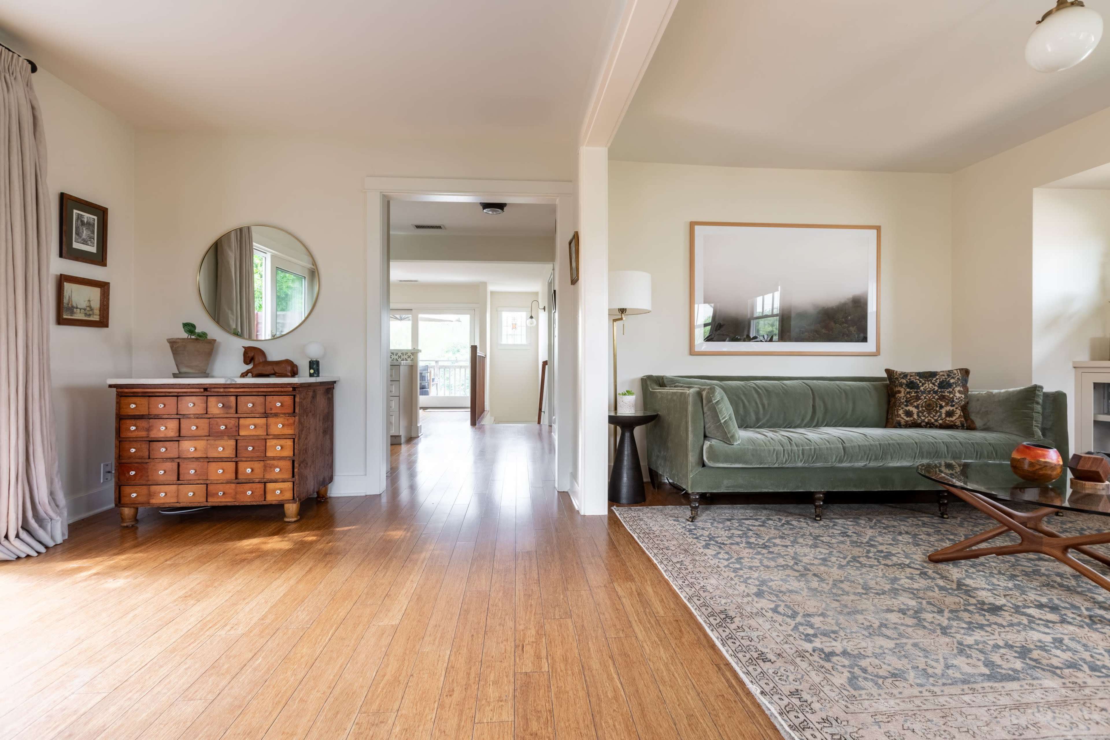 The image shows a living room with a green velvet sofa, a wooden side table, a decorative rug, and natural light coming through windows.