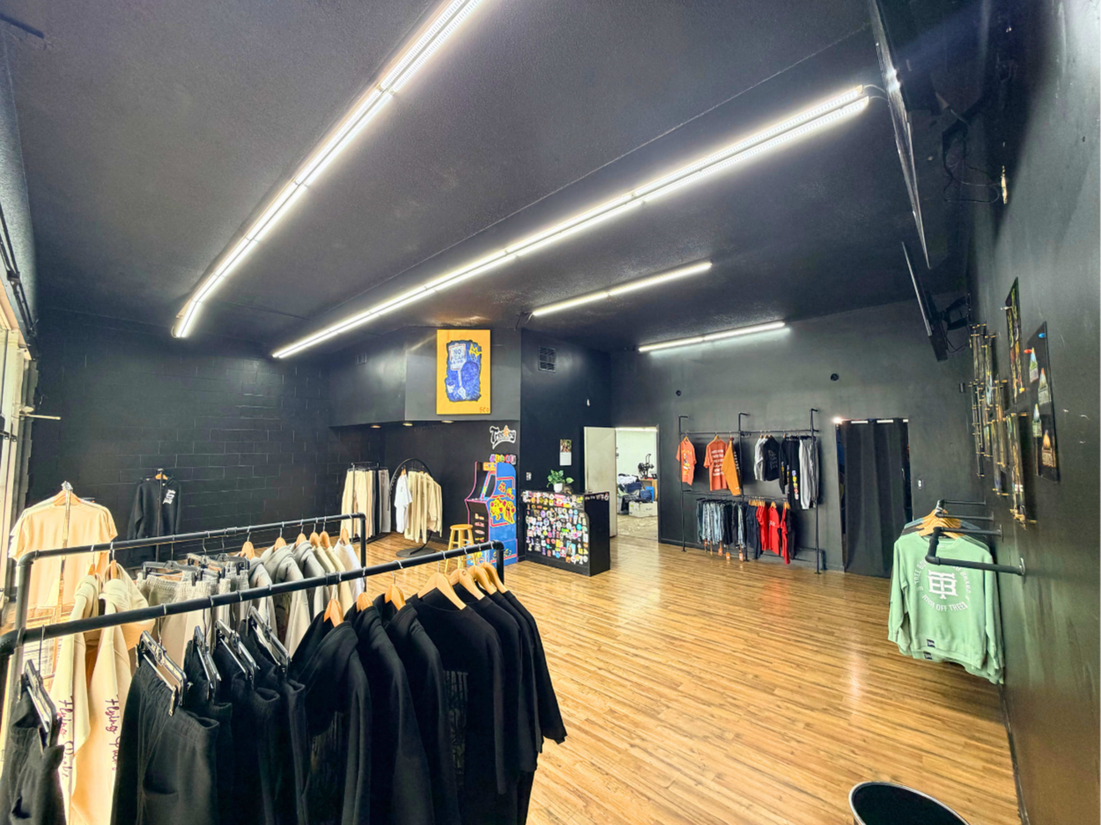 The image shows the interior of a clothing store with racks of apparel and a black wall backdrop, featuring overhead lighting and a framed picture on the wall.