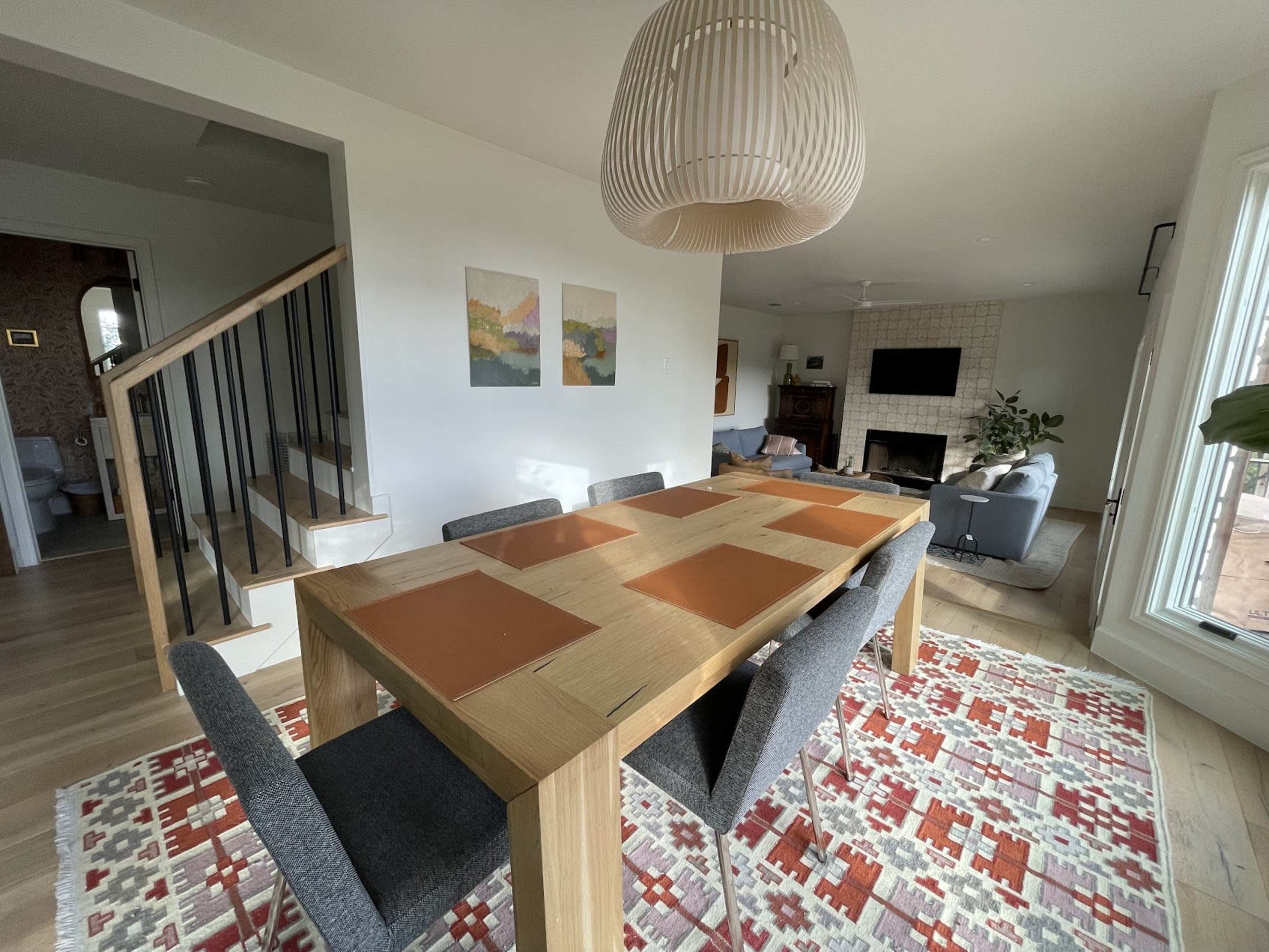 The image shows a dining area with a wooden table and gray chairs on a patterned rug, accompanied by a staircase and a fireplace in the background.