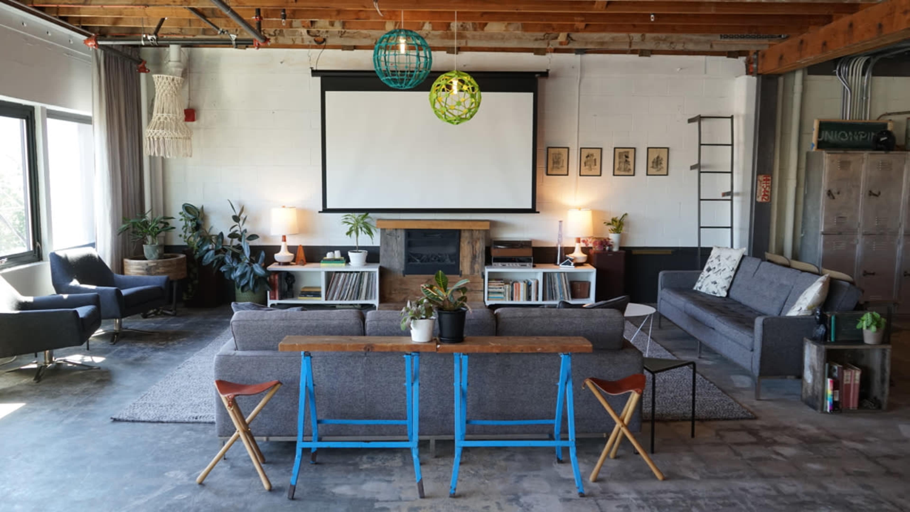 A contemporary living area features two gray sofas, a wooden coffee table, a large screen for projection, and various plants against a backdrop of exposed wood beams and white walls.