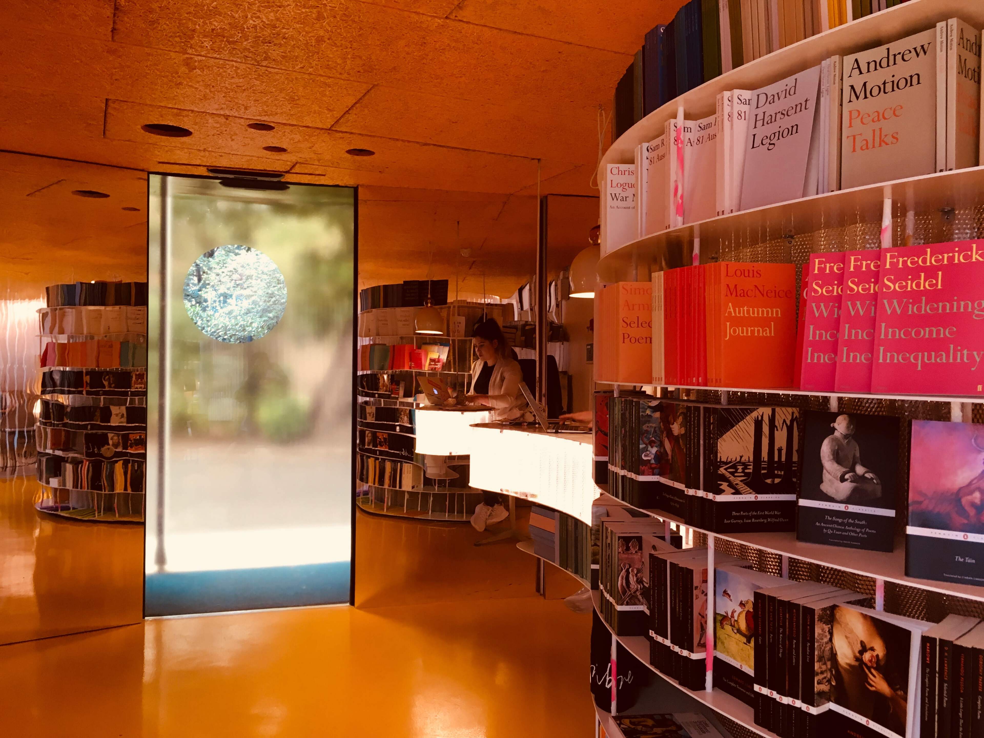 The image shows a modern library setup with shelves of books arranged in circular patterns and a person standing at a desk near a large, round glass door.