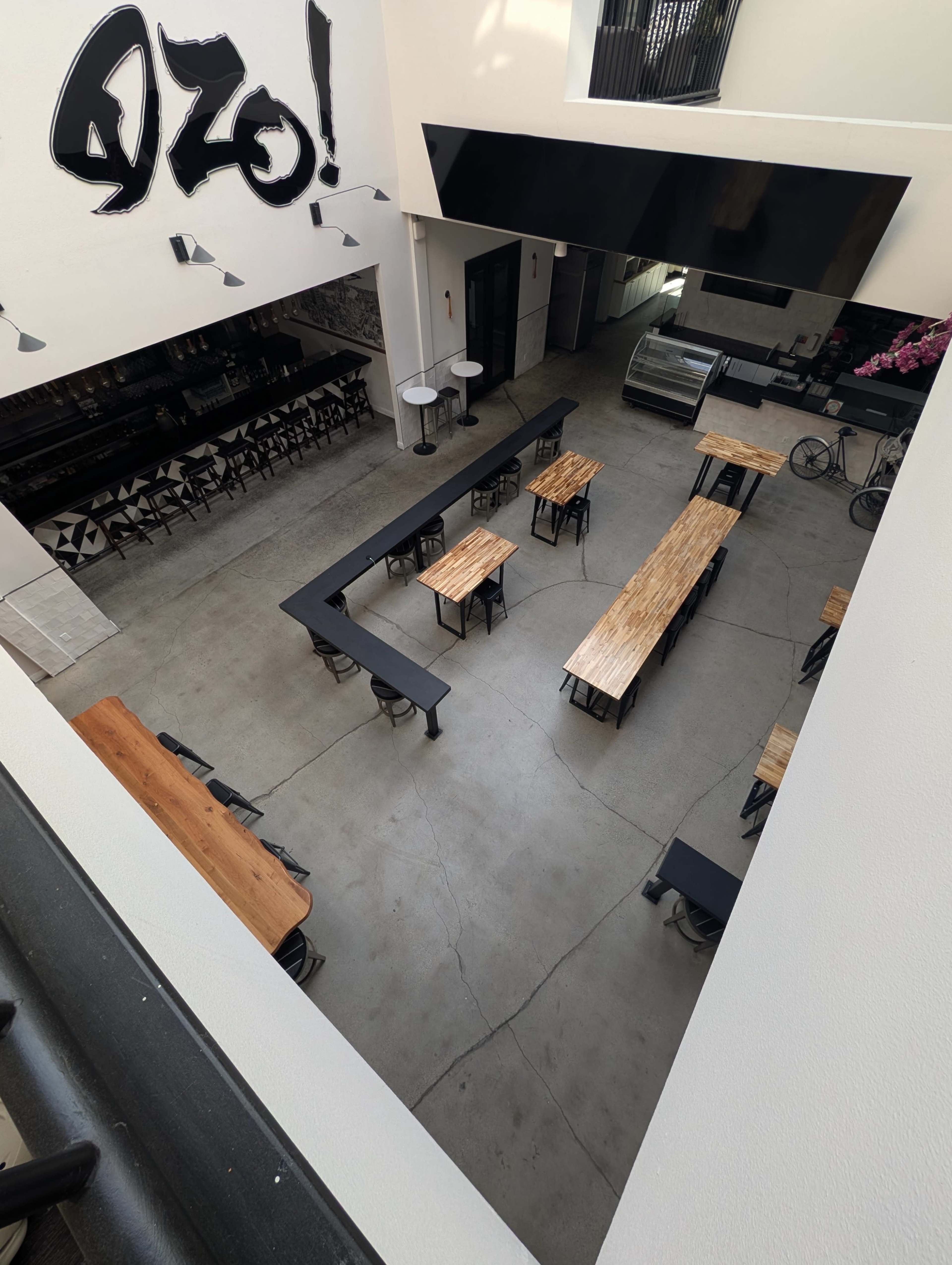 The image shows a spacious indoor café or restaurant with several long wooden tables, black metal chairs, and a counter area in a minimalist design.
