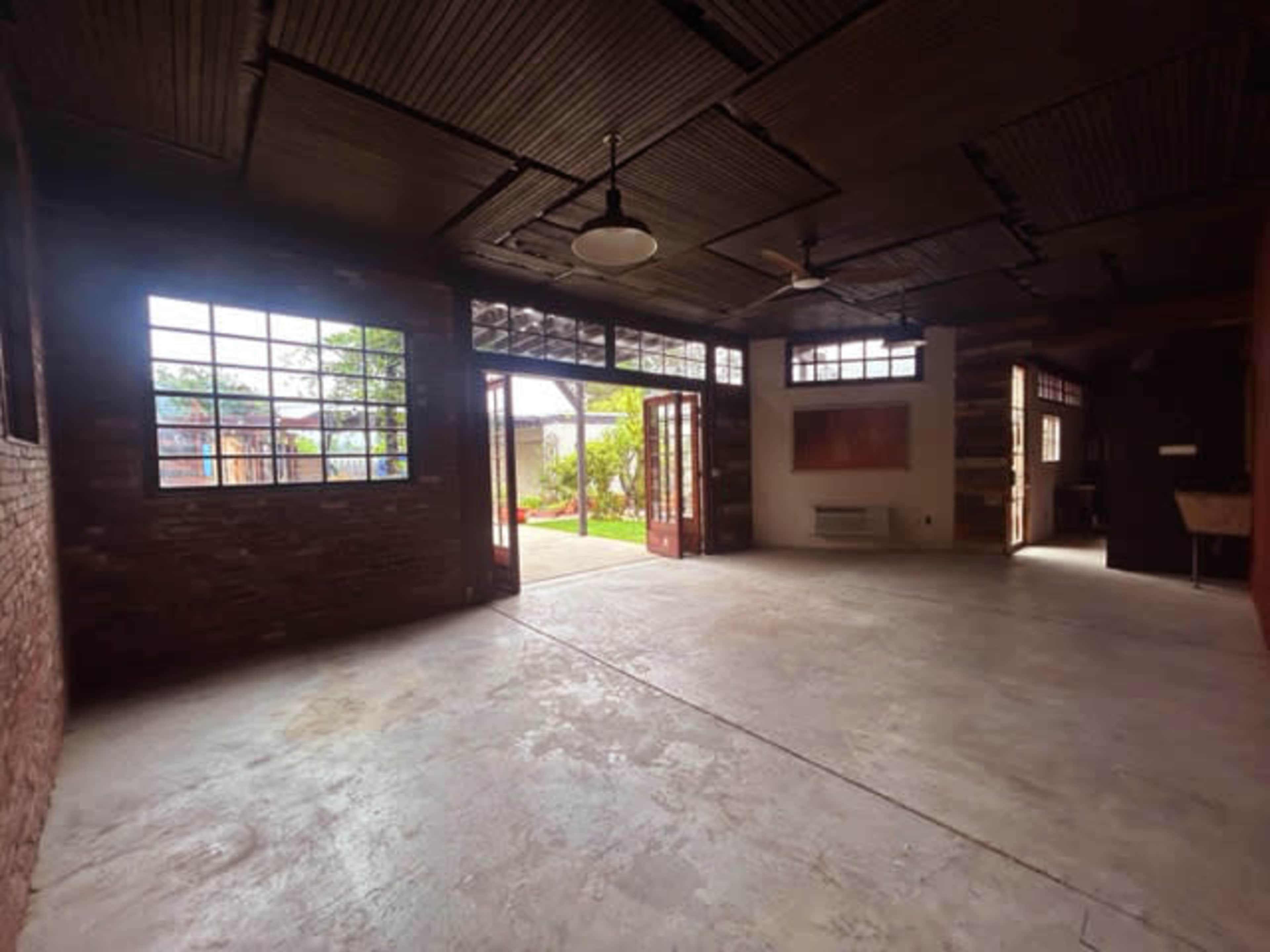 A spacious room with large windows, double doors, and an industrial-style ceiling, leading to a garden area outside.