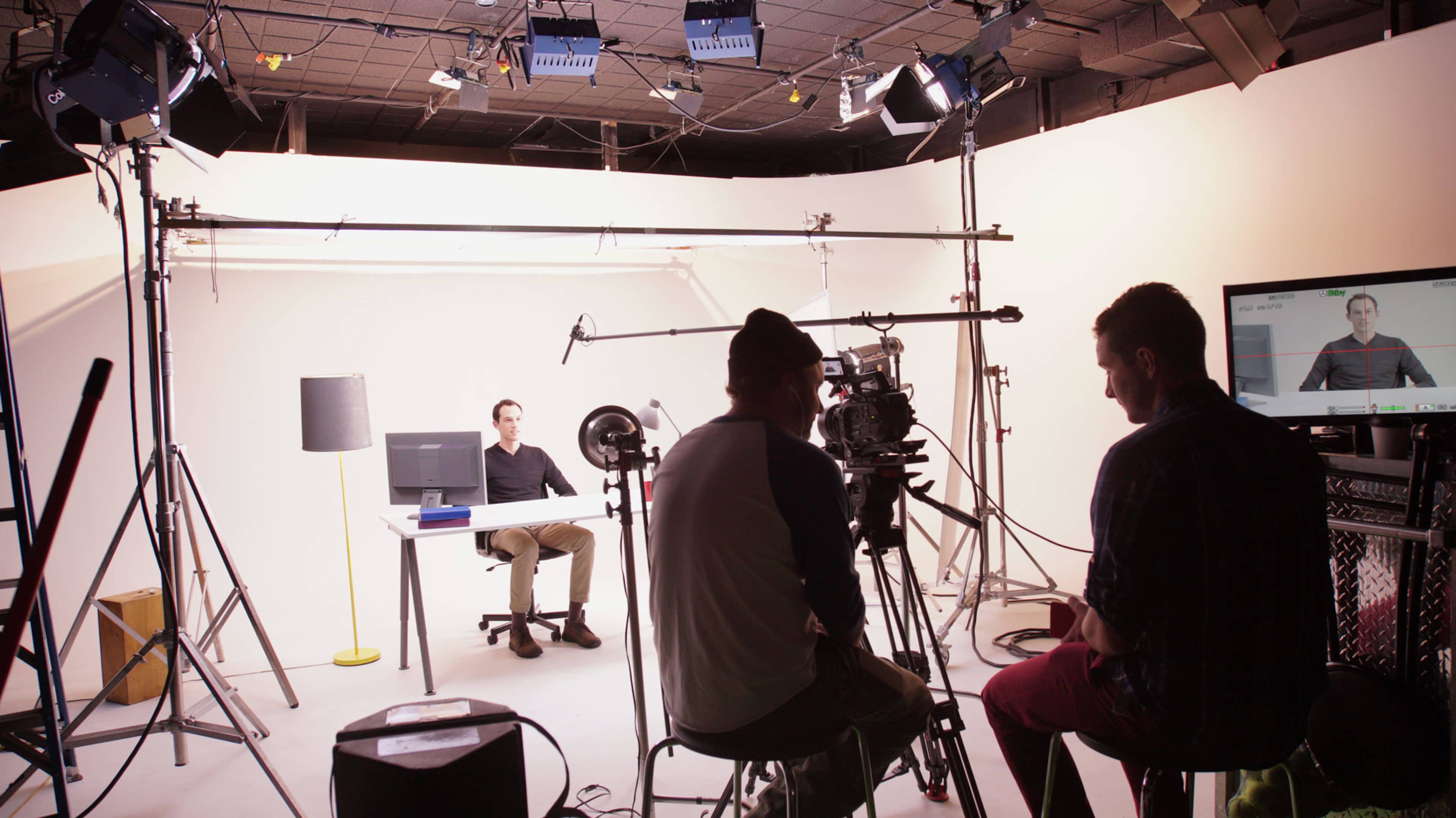 A video production crew sets up equipment in a studio where a person is seated at a desk.