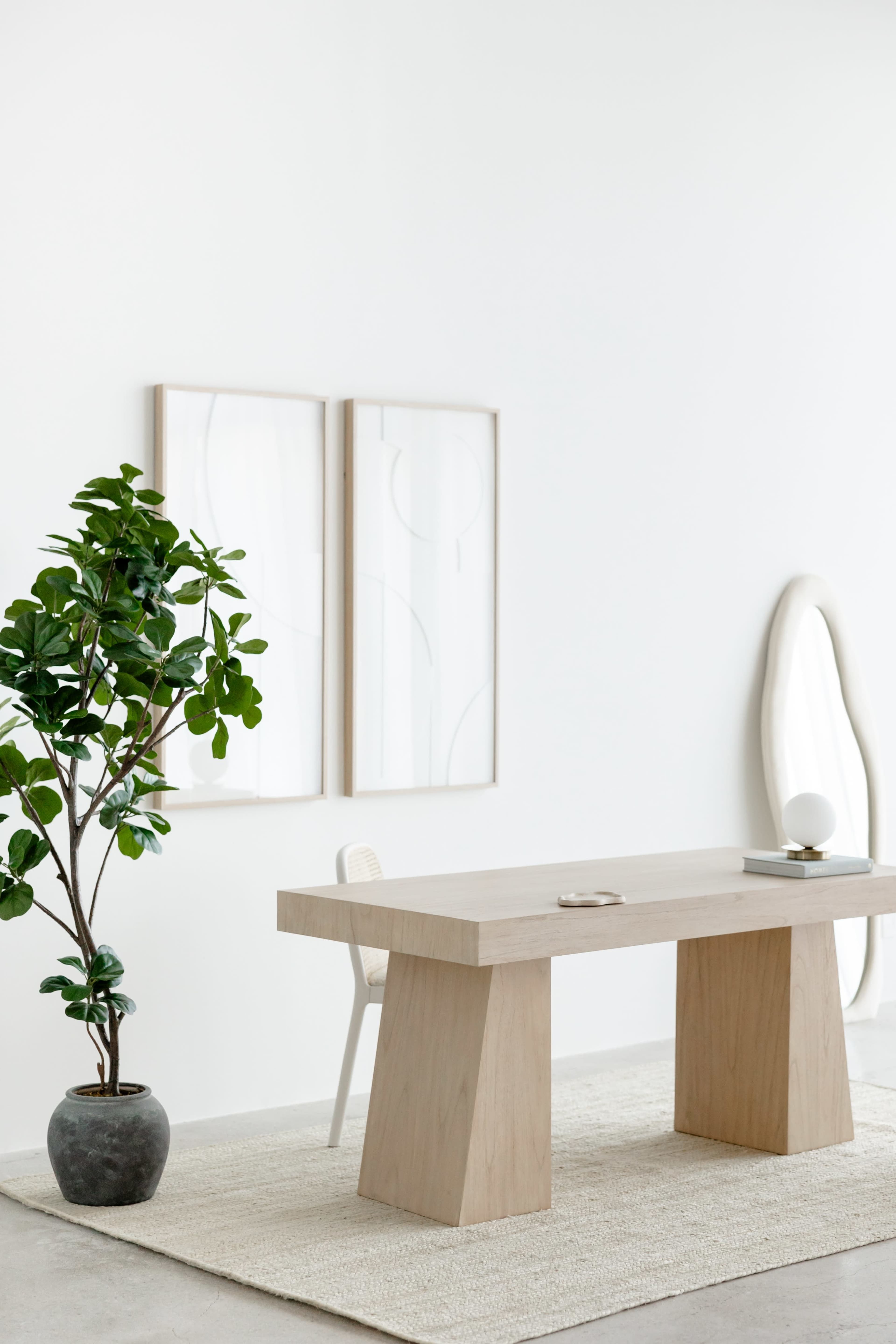 A minimalist workspace features a light wood table, a light chair, a potted plant, and abstract art on the wall.