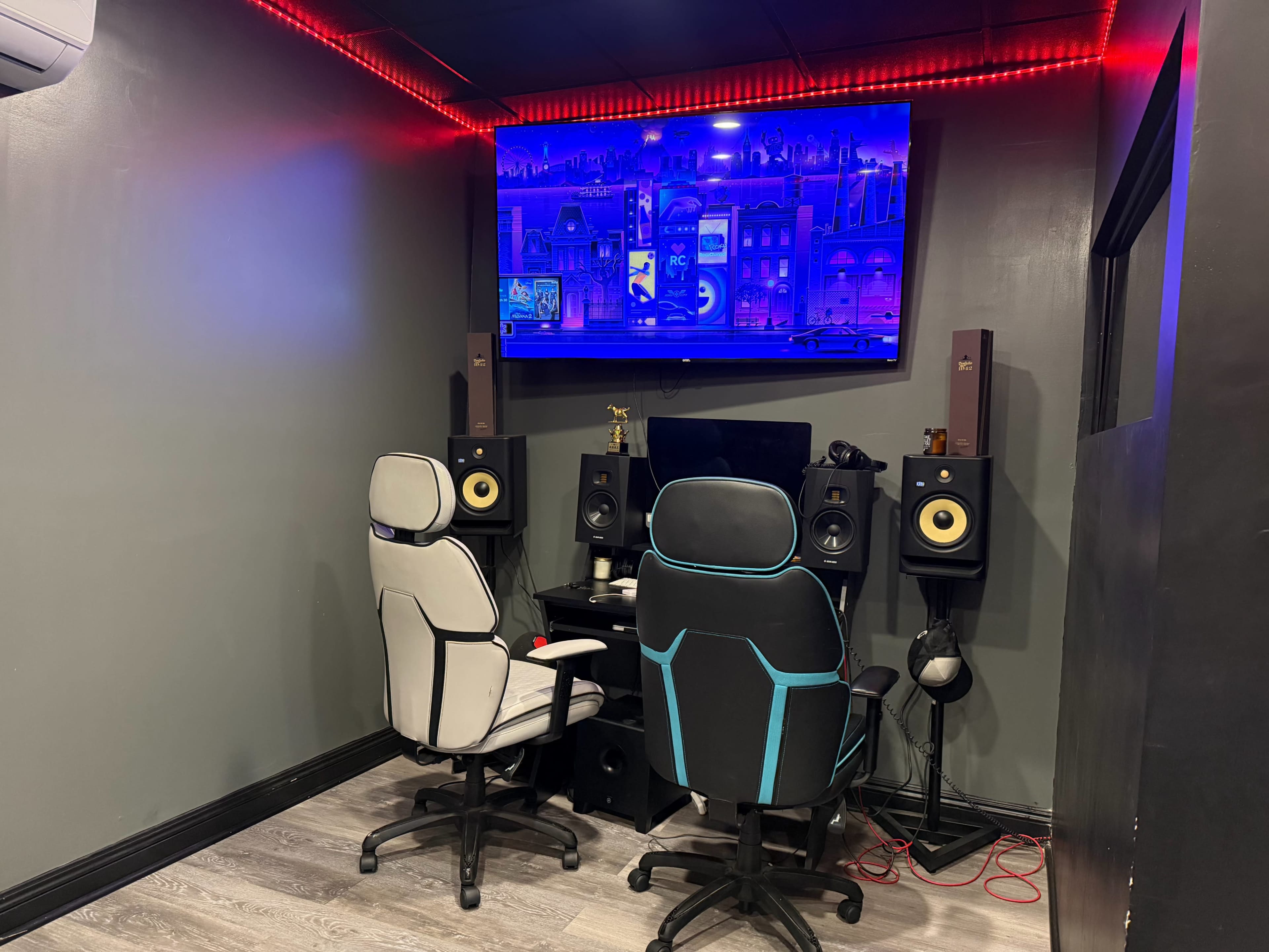 The image shows a small recording or gaming studio featuring two ergonomic chairs, a desk with a computer monitor, and speakers mounted on either side, all set against a dimly lit gray wall with red accent lighting.