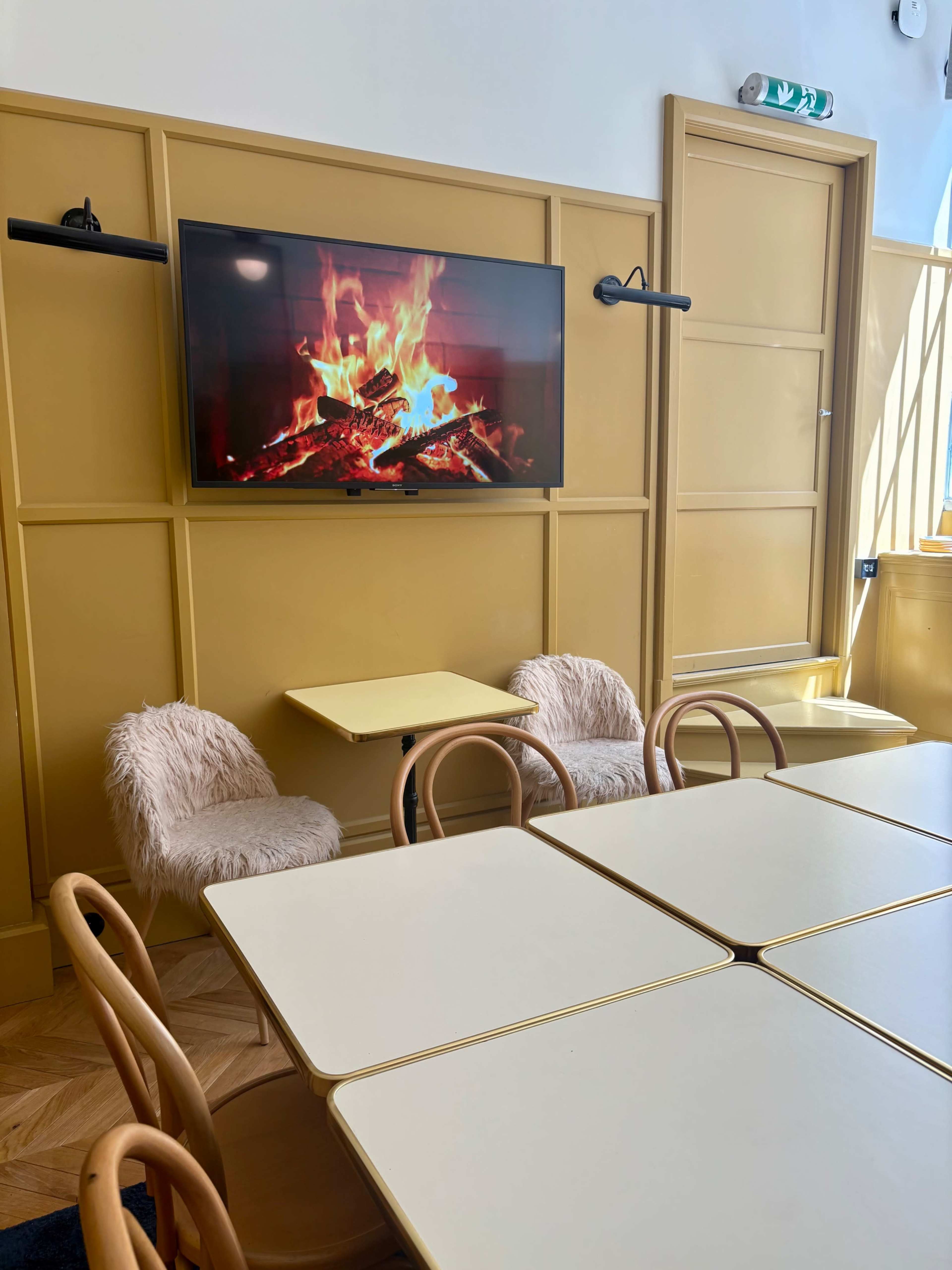 The image shows a cozy café interior featuring light-colored tables, wooden chairs, and a wall-mounted television displaying a flickering fireplace.