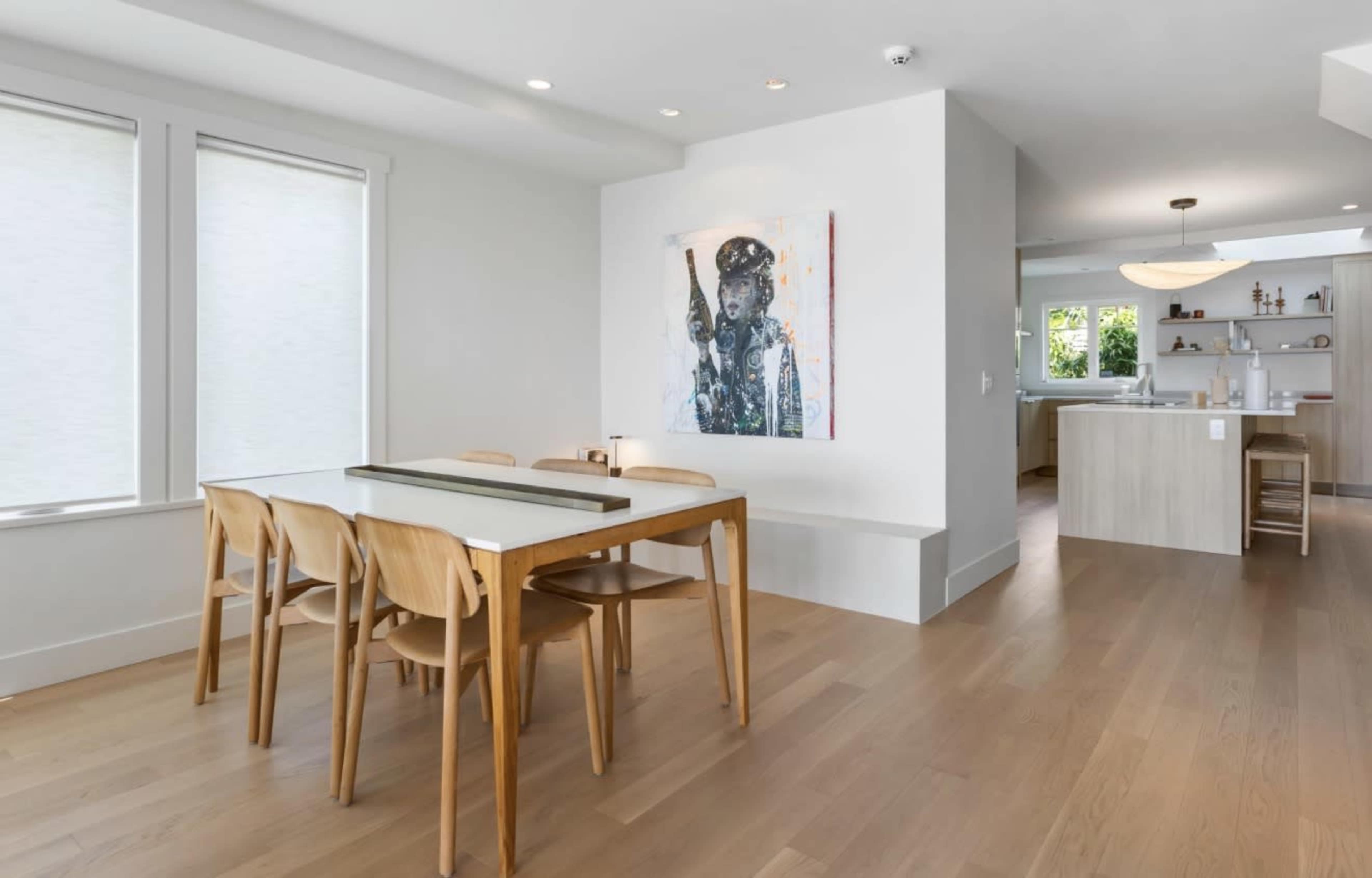 The image features a modern dining area with a wooden table and six chairs, adjacent to a small kitchen in a well-lit space.