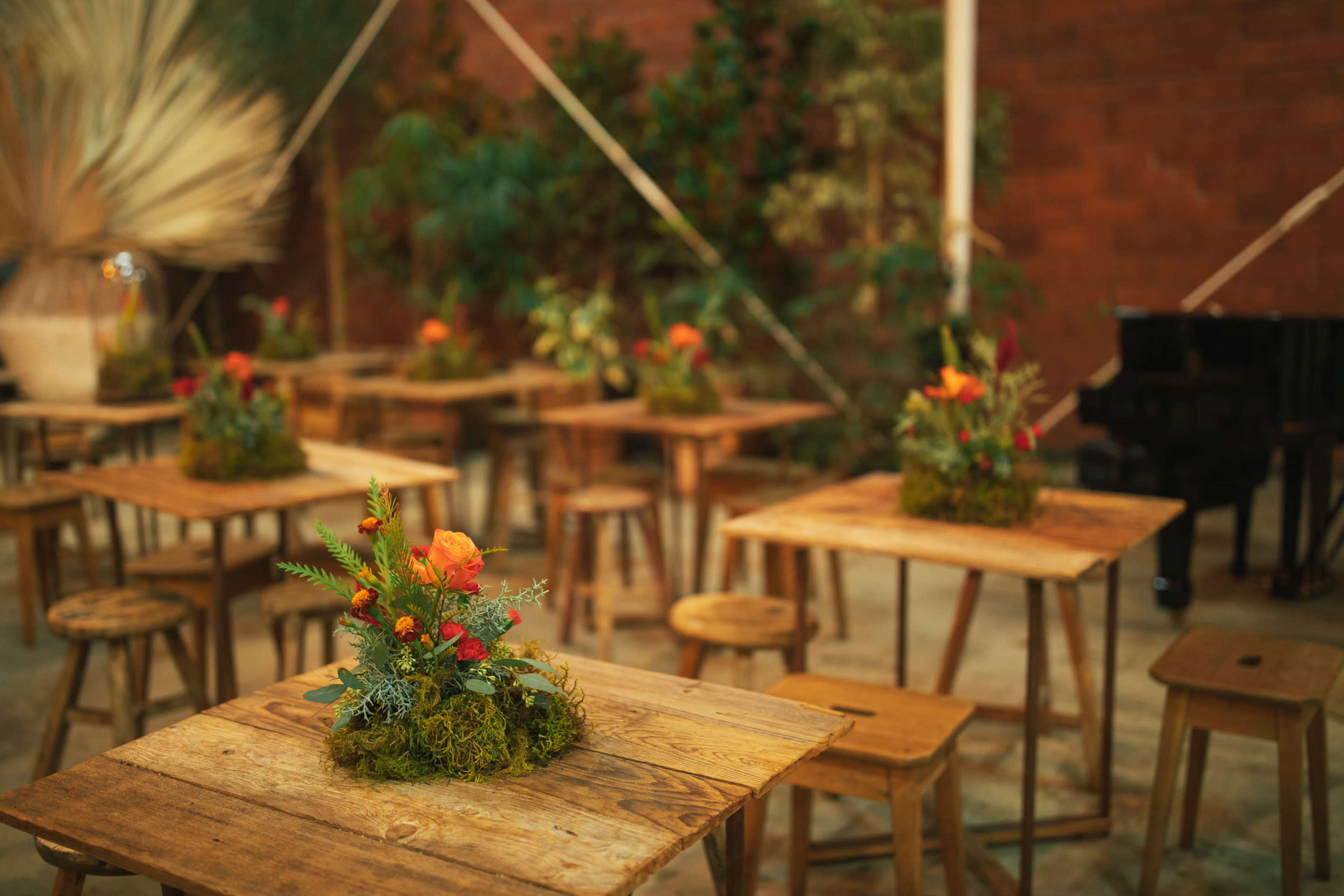 The scene features wooden tables adorned with floral arrangements in moss, surrounded by stools and a black piano in a decorated indoor space.