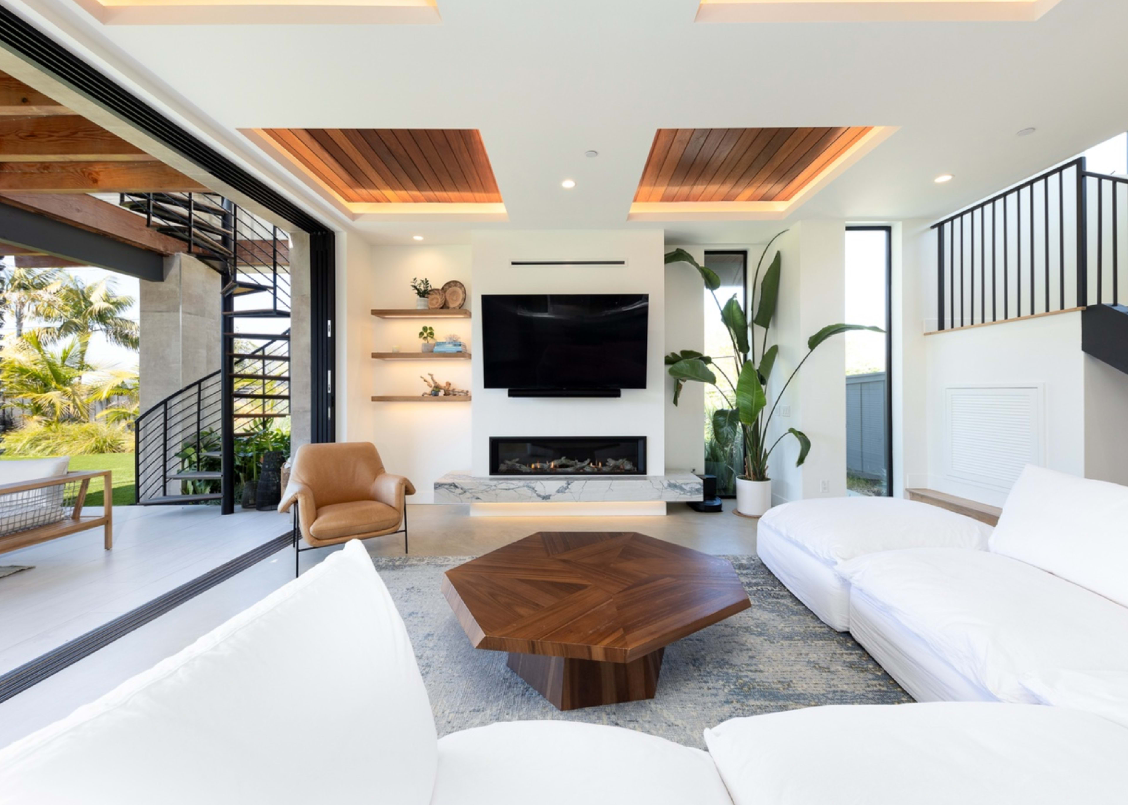 A modern living room with a white sectional sofa, a wooden coffee table, a wall-mounted television, and large windows overlooking a patio with greenery.