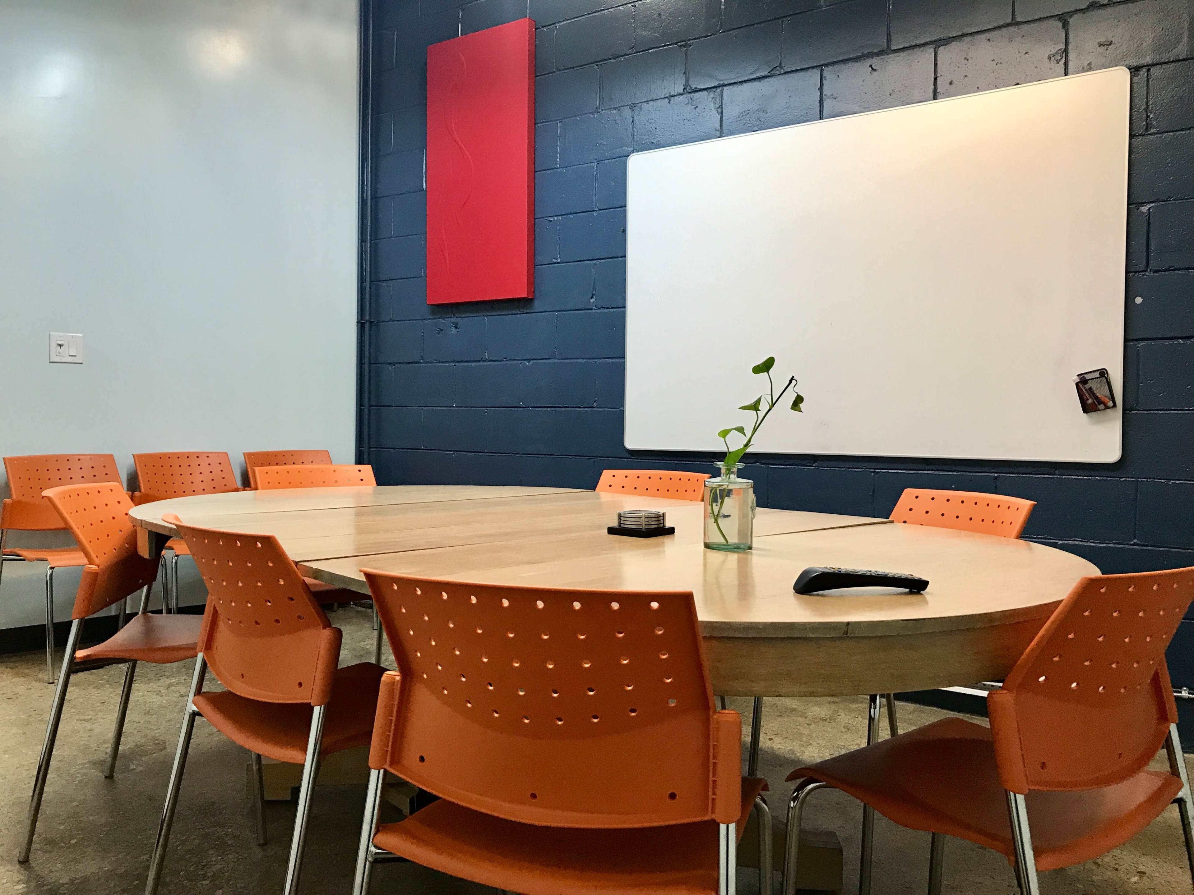 A round wooden table surrounded by eight orange chairs is set in a room with a blue wall and a whiteboard.