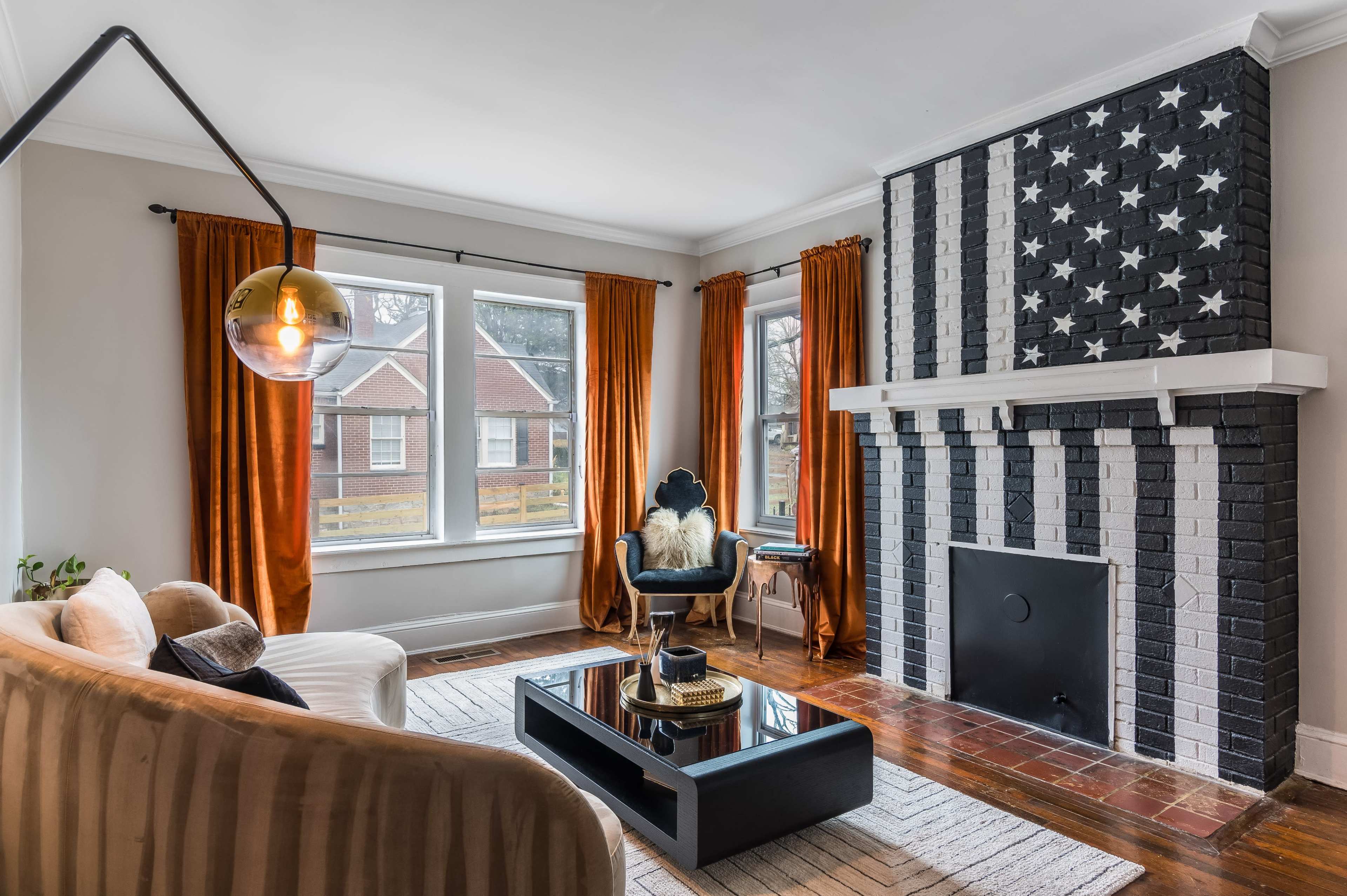 The living room features a striped black and white fireplace, large windows draped in orange curtains, and modern furniture arranged around a coffee table.