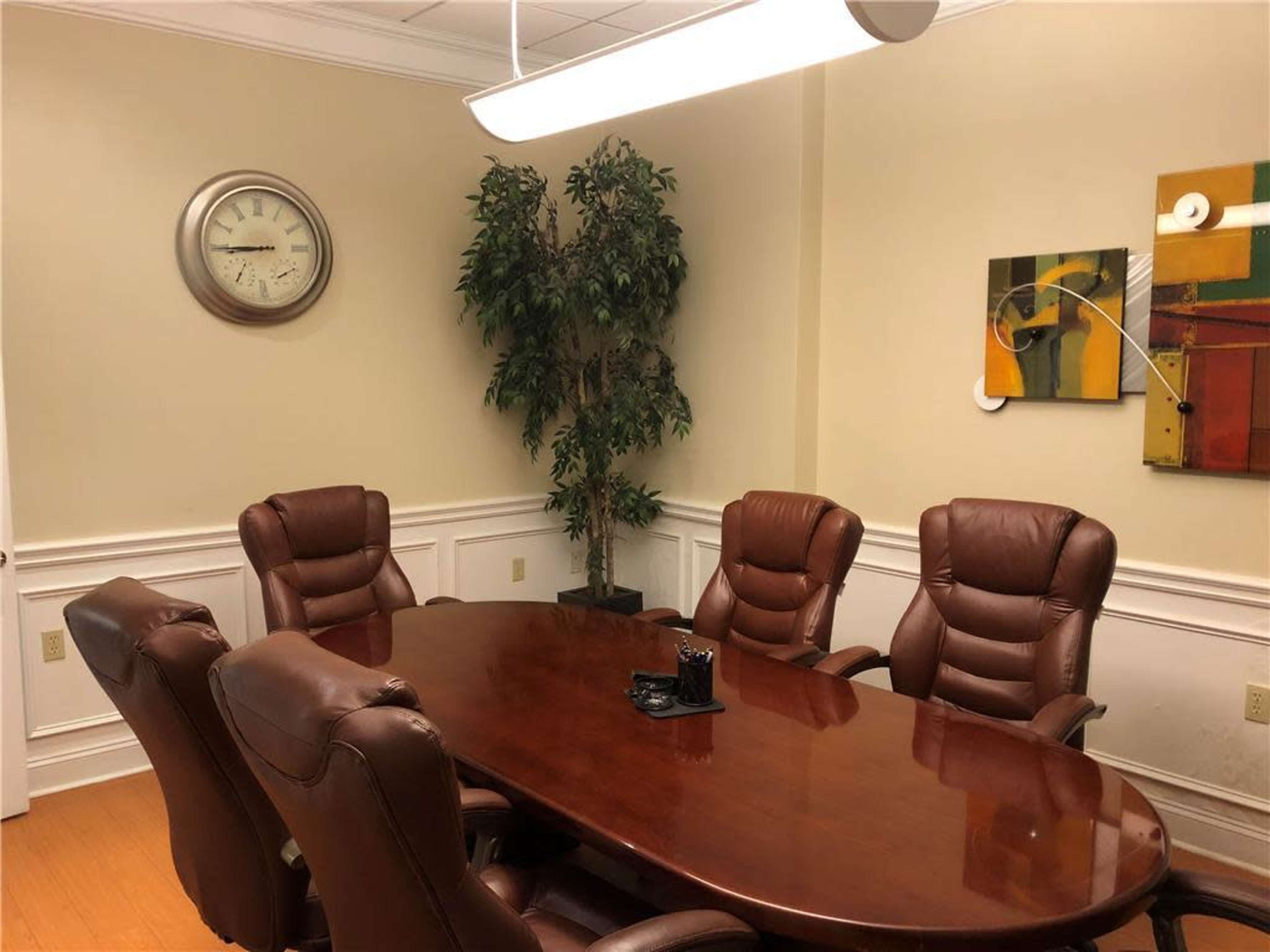 The image shows a conference room featuring a large wooden table surrounded by five leather chairs, with a clock and artwork on the walls.