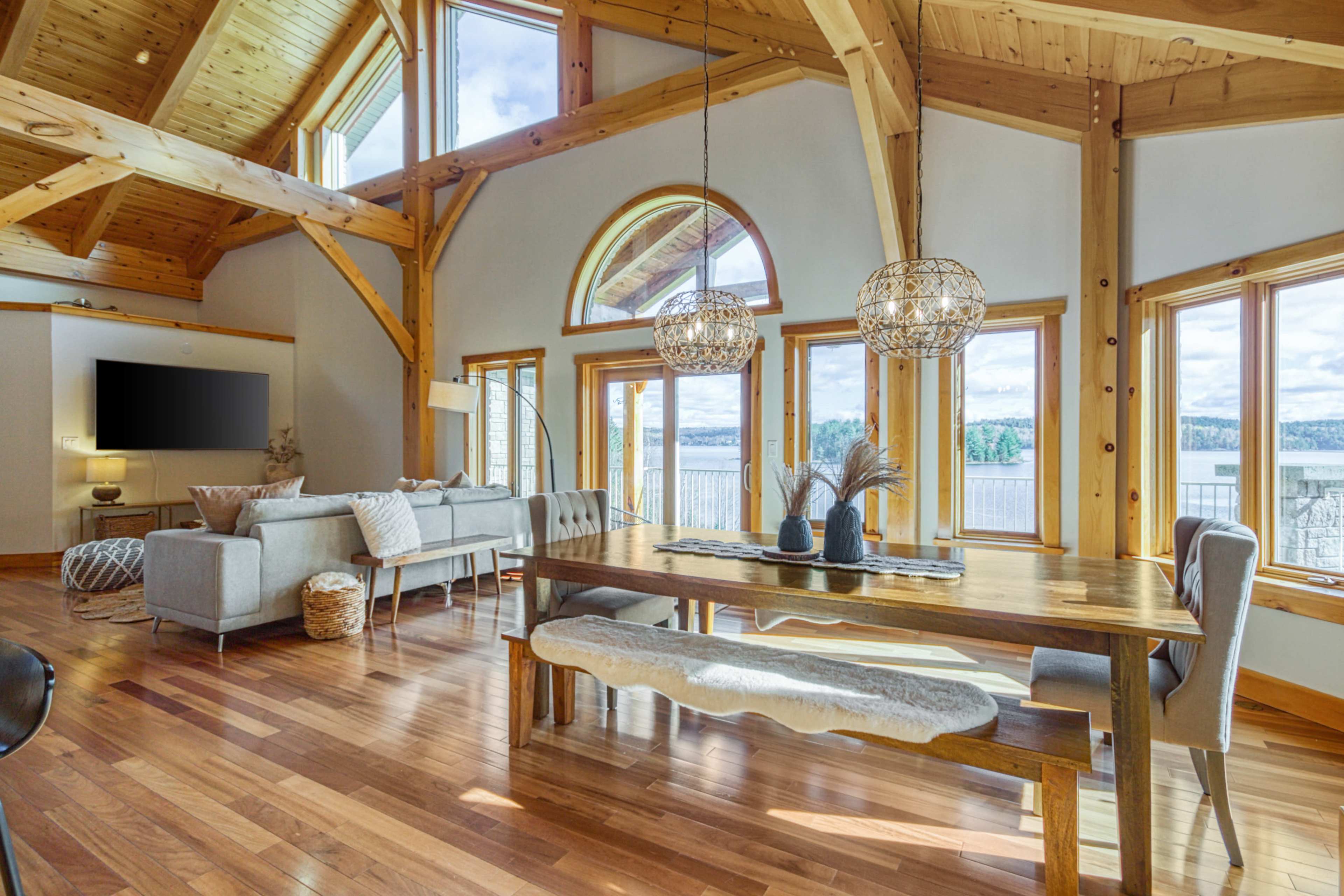 A bright, spacious interior features wooden beams, large windows overlooking a body of water, and a dining table surrounded by comfortable seating.