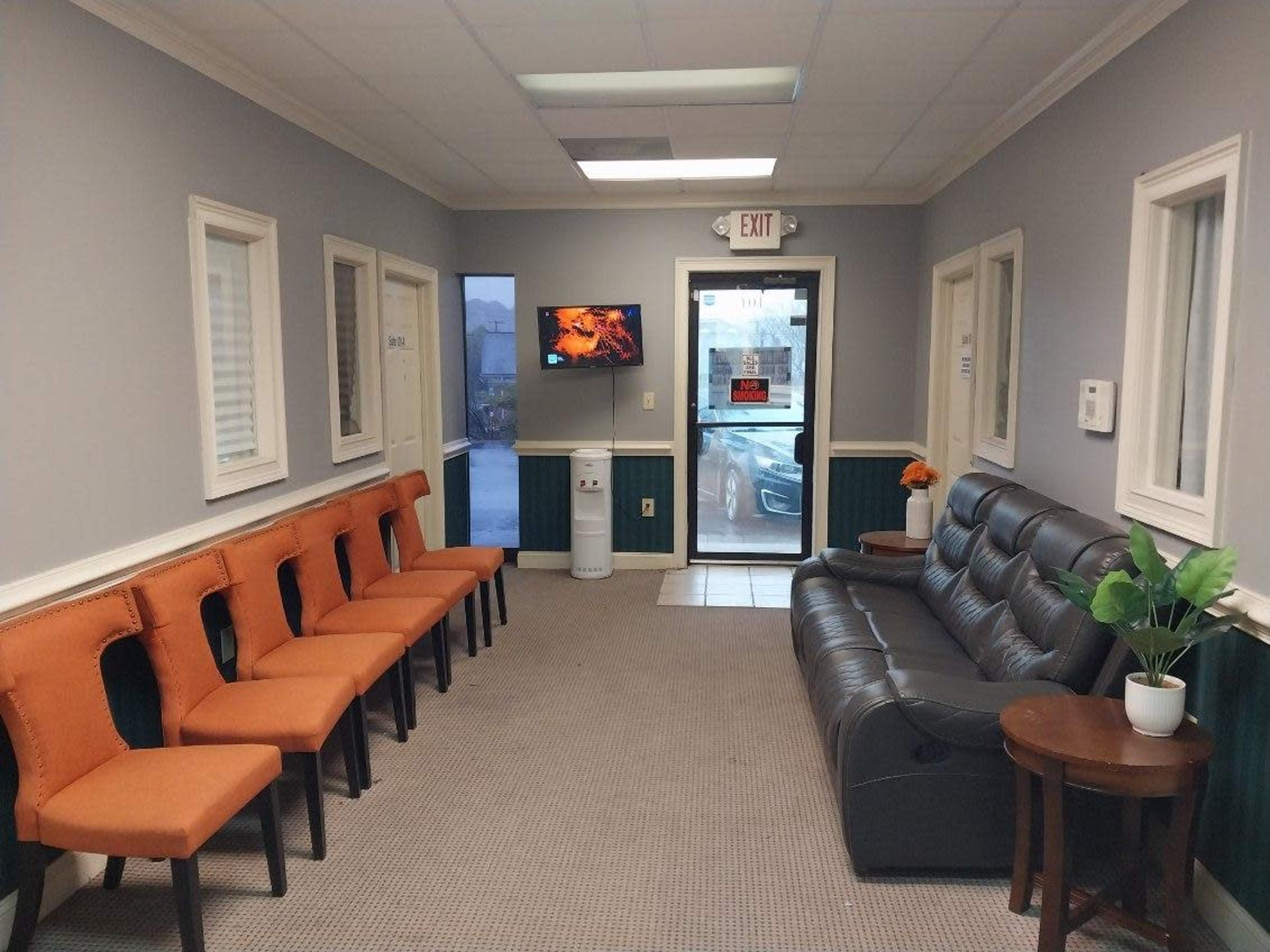 A waiting room with several orange chairs along one wall, a black sofa, and a TV mounted on the opposite wall near a glass door.