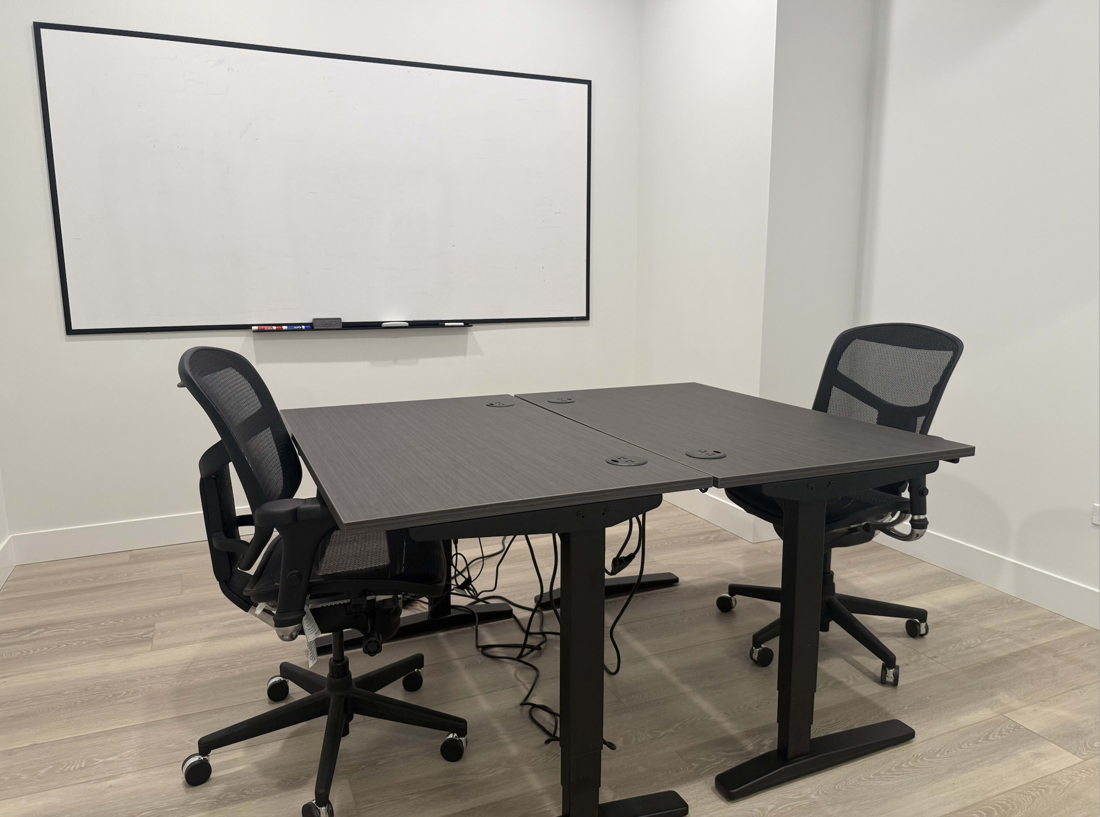 A small meeting room features a split conference table with two adjustable chairs and a blank whiteboard in the background.