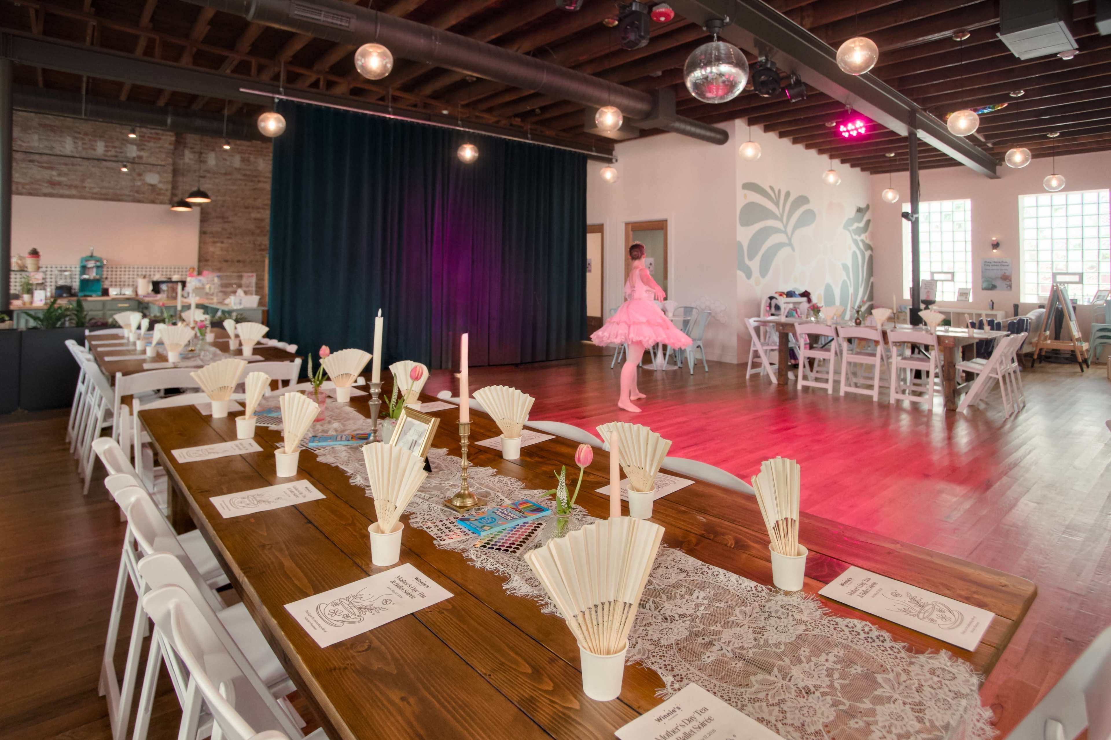 A spacious event venue features a long wooden dining table set with paper fans and candles, while a performer dances in the background under colorful lighting.