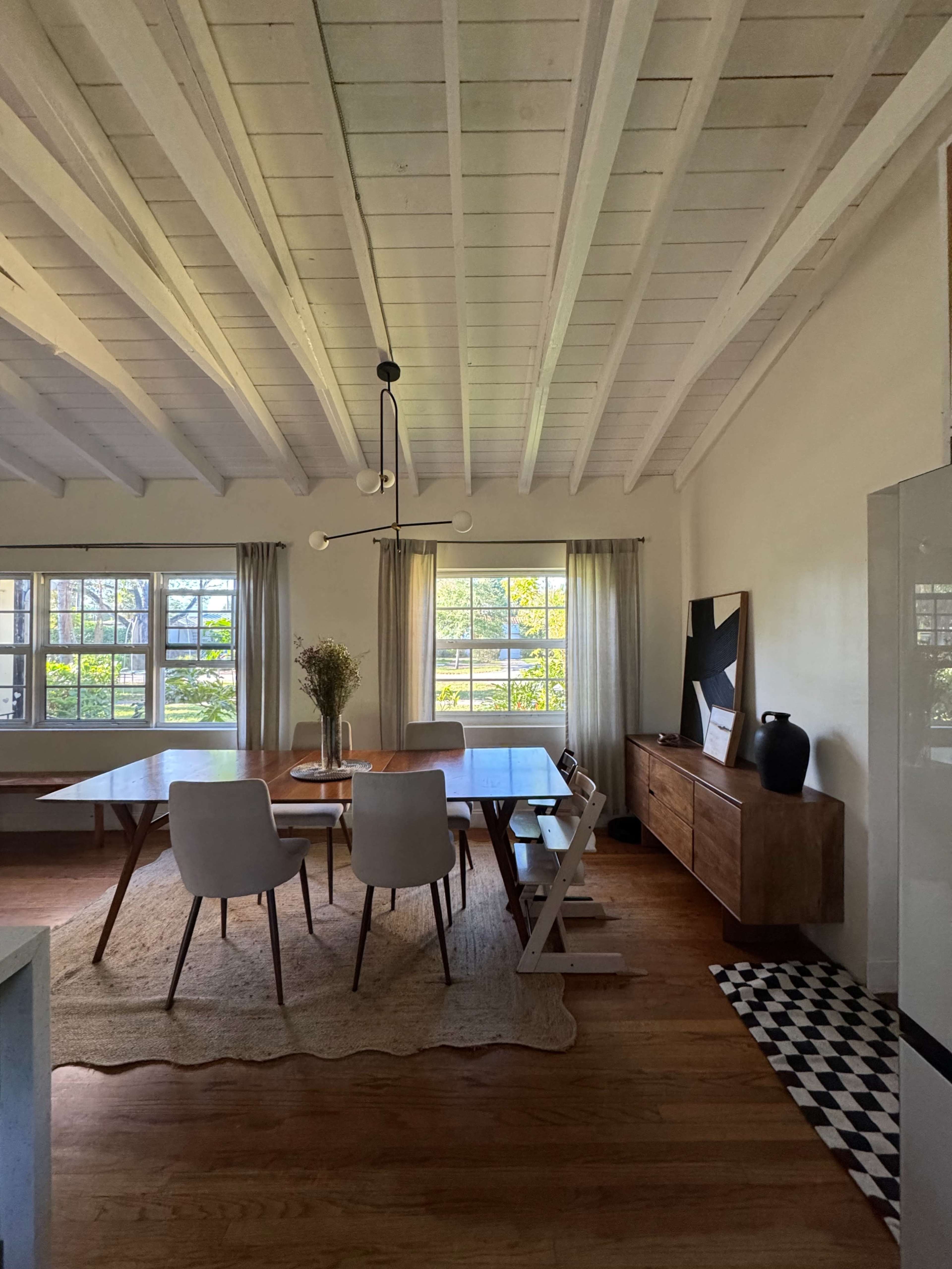 The image shows a bright dining area with a wooden table surrounded by white chairs, a sideboard, and large windows letting in natural light.