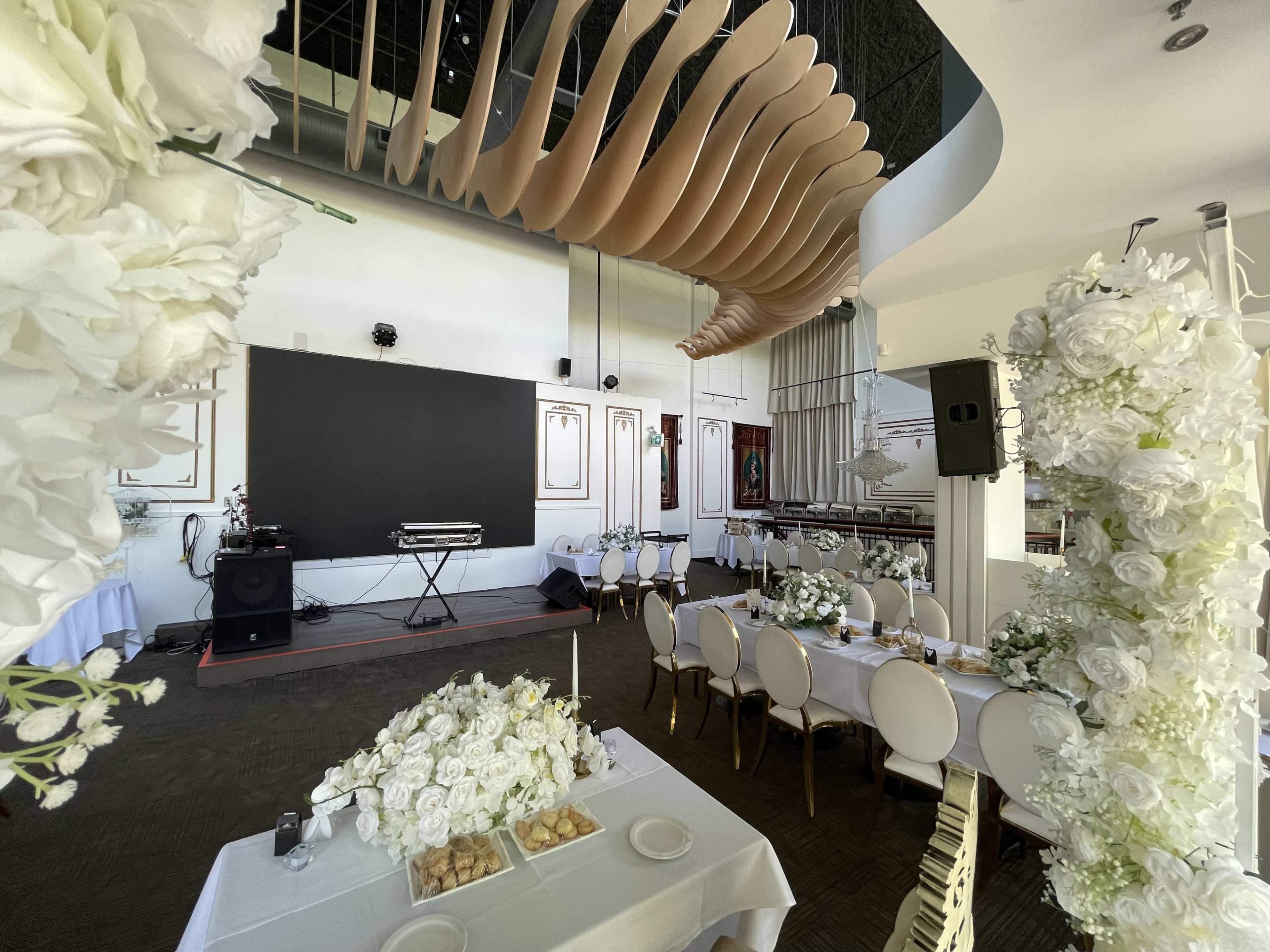 The image shows a spacious event venue set up for a gathering, featuring tables adorned with white floral centerpieces and a stage area with sound equipment.