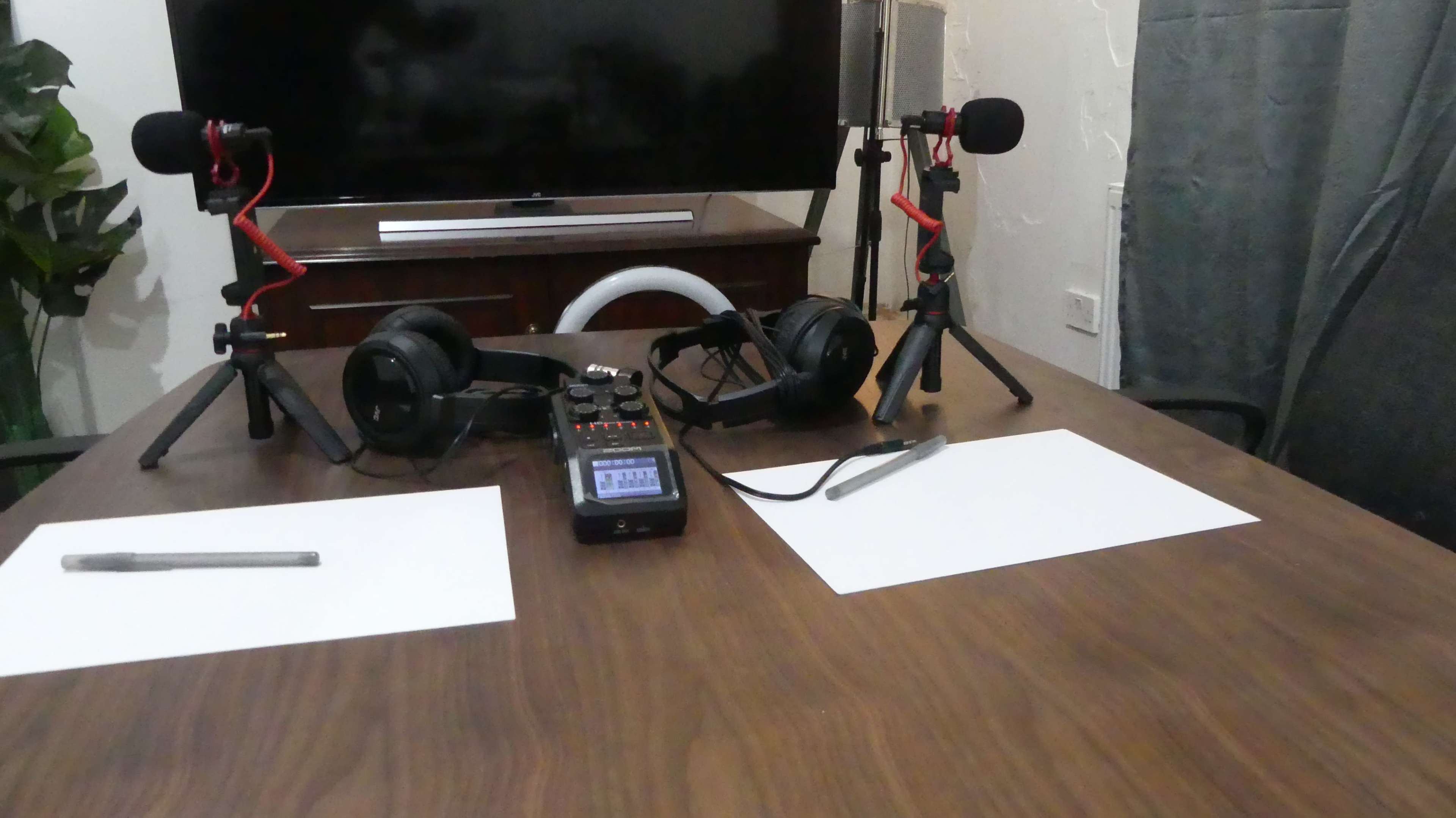 A wooden table is set up with two microphones on stands, headphones, a recording device, and blank sheets of paper on each side.