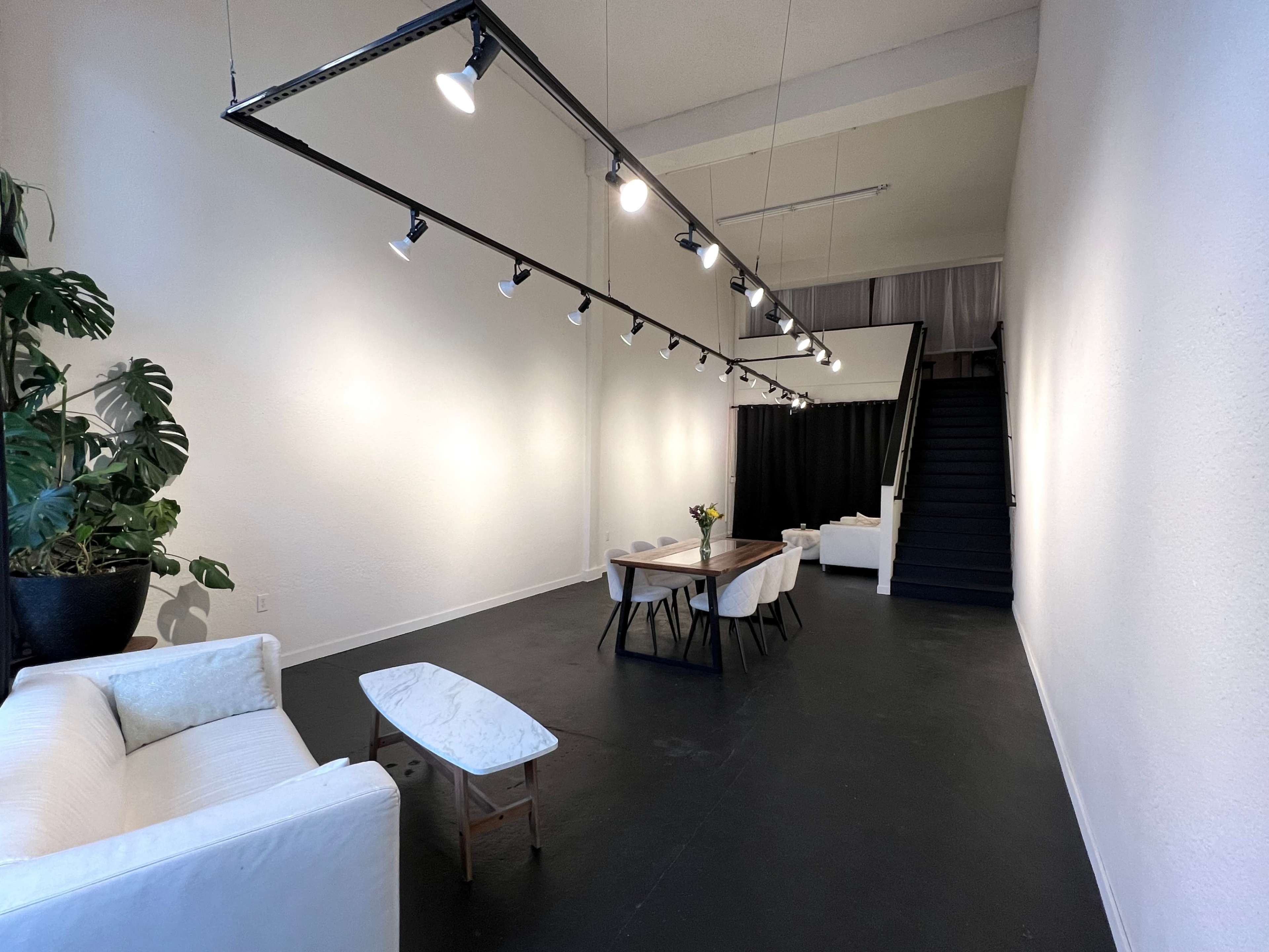 A spacious, well-lit interior featuring a seating area with white couches, a dining table, and a staircase leading to an upper level.