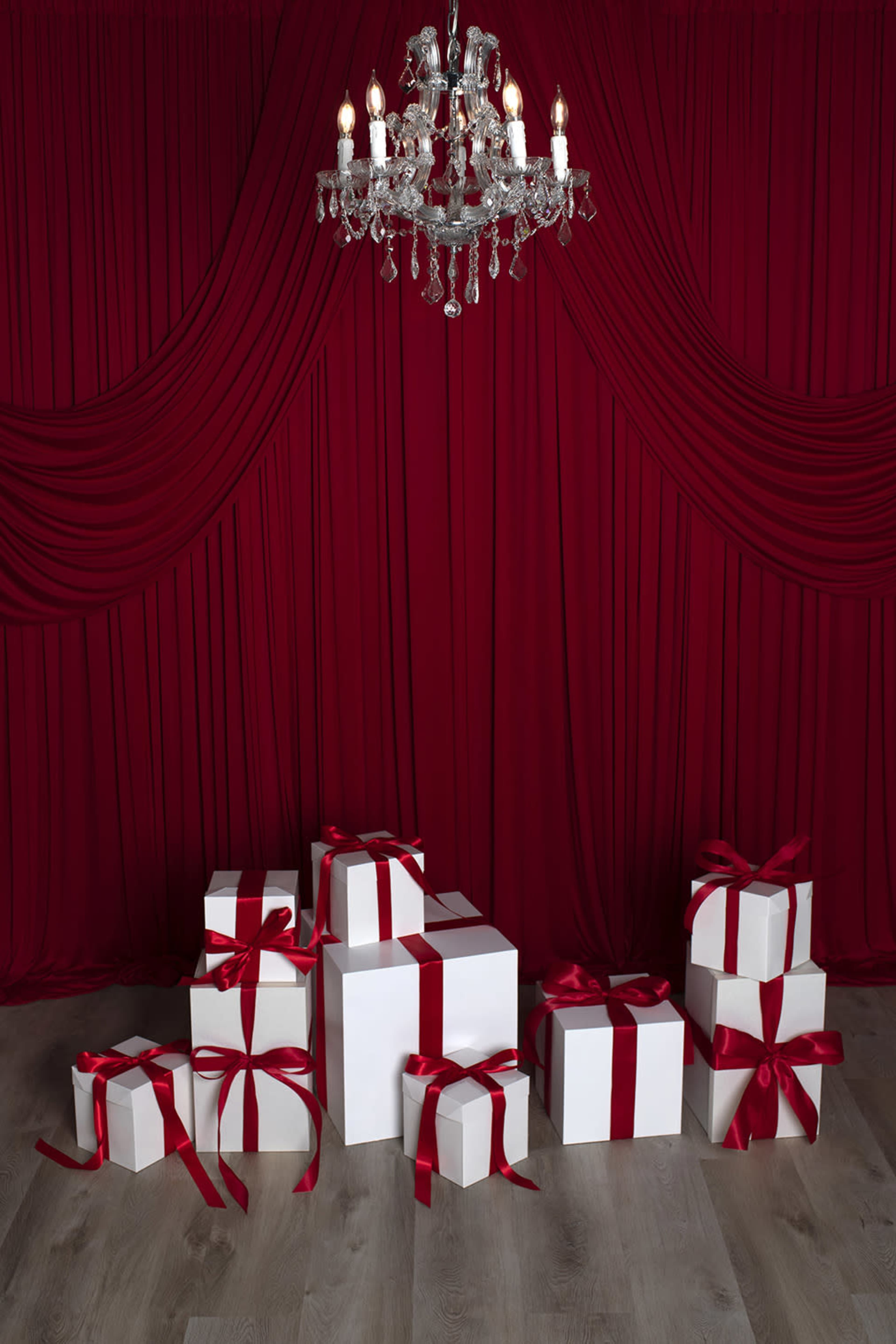 A collection of white gift boxes with red ribbons is arranged on a wooden floor in front of a red draped backdrop and a crystal chandelier.