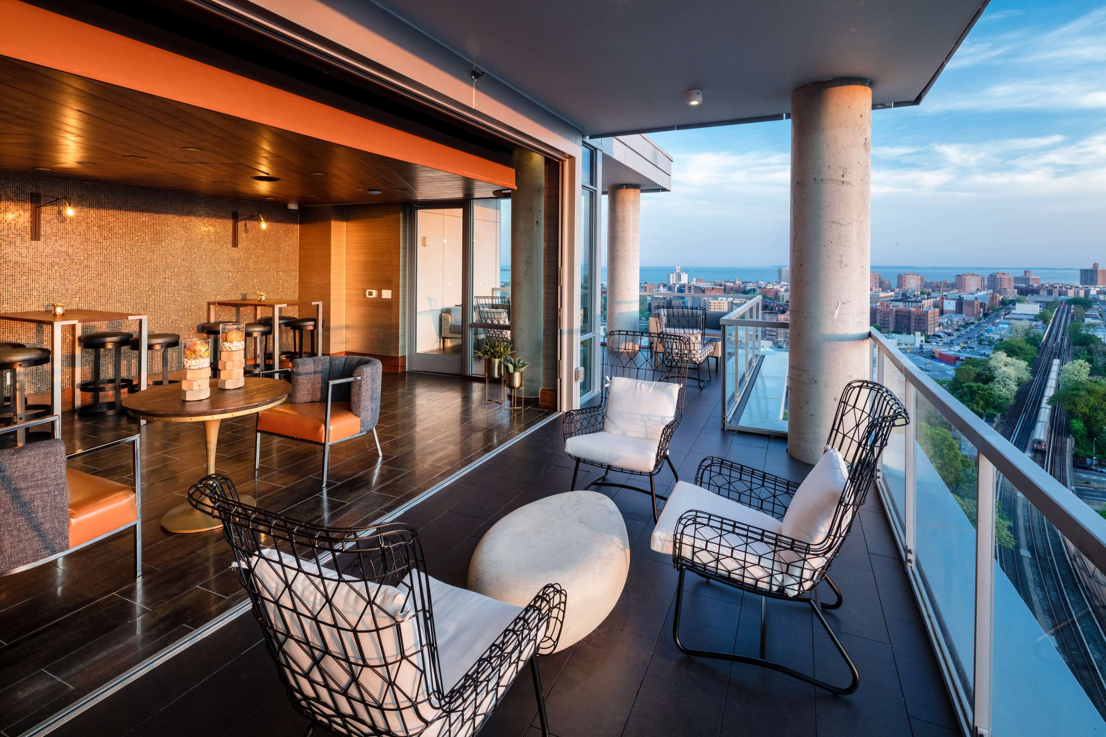 A modern balcony features wire chairs, a circular table, and a view of the city skyline and train tracks.
