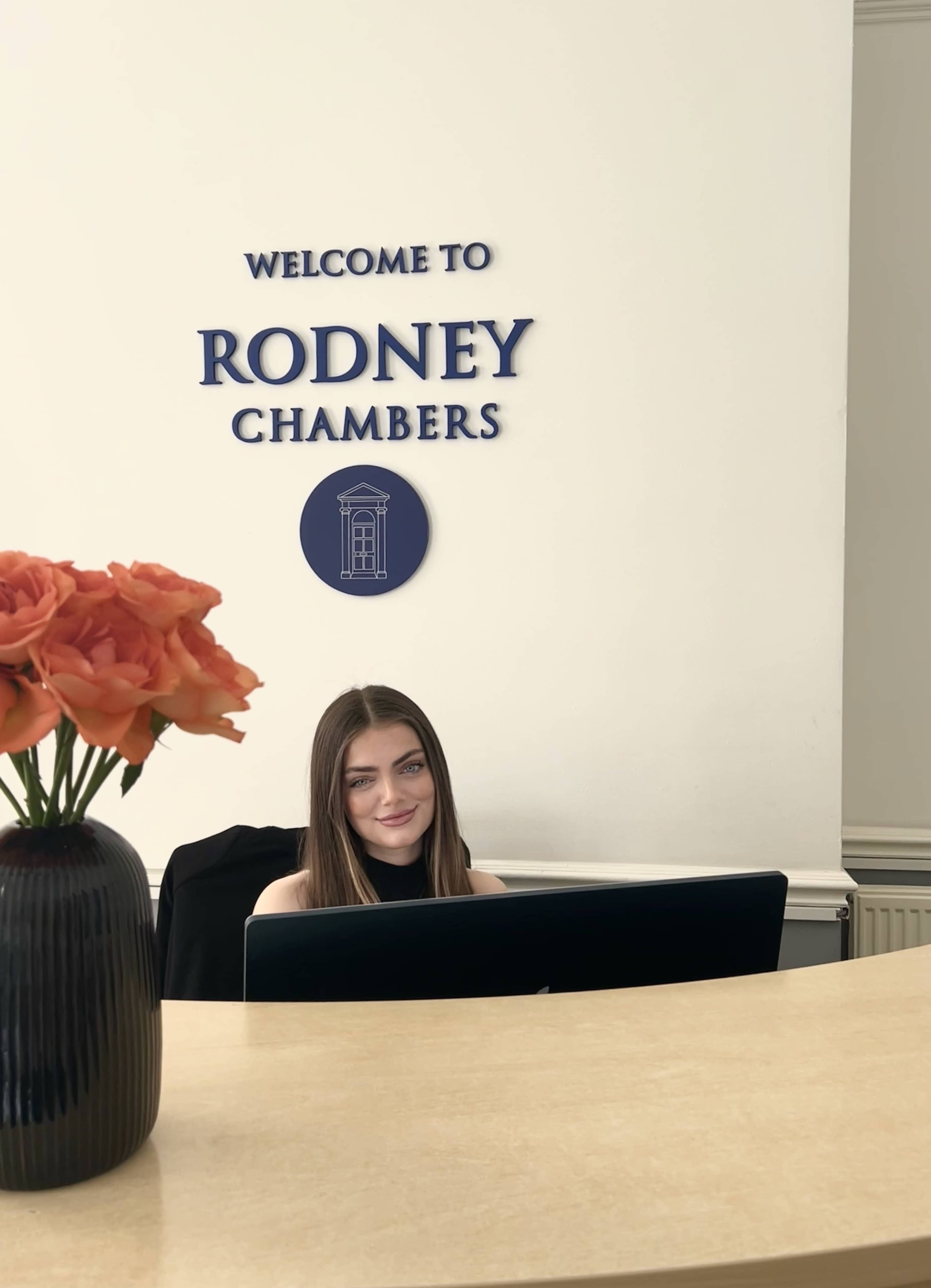 A young woman sits at a reception desk in front of a wall sign that reads "Welcome to Rodney Chambers," with a vase of orange flowers nearby.