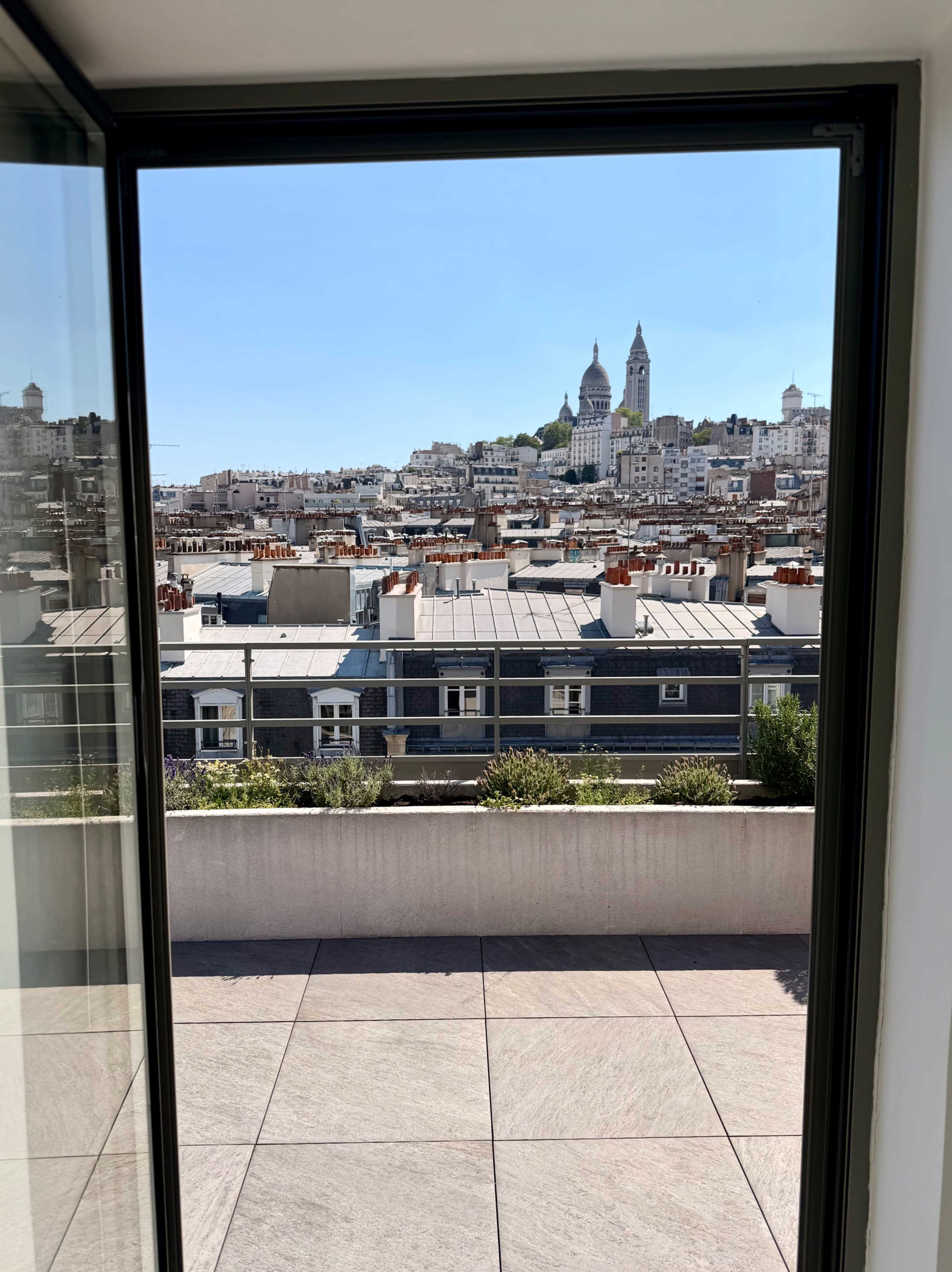 The image shows a view from an open door with a terrace, overlooking rooftops and the cityscape of Paris, including the silhouette of a prominent church or basilica in the background.