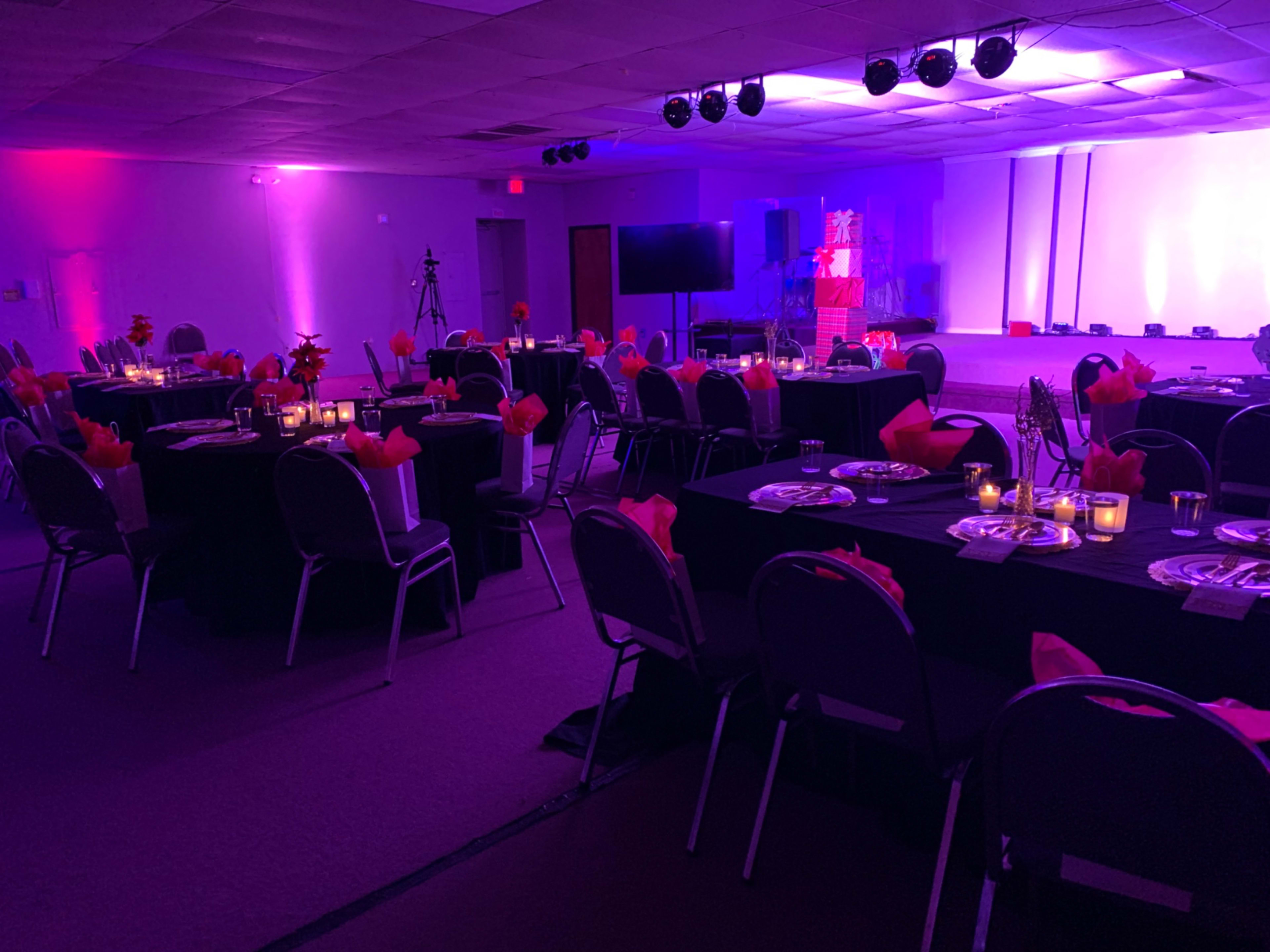A banquet hall set up with tables covered in black cloth, adorned with pink napkins and lit candles, all under purple accent lighting.