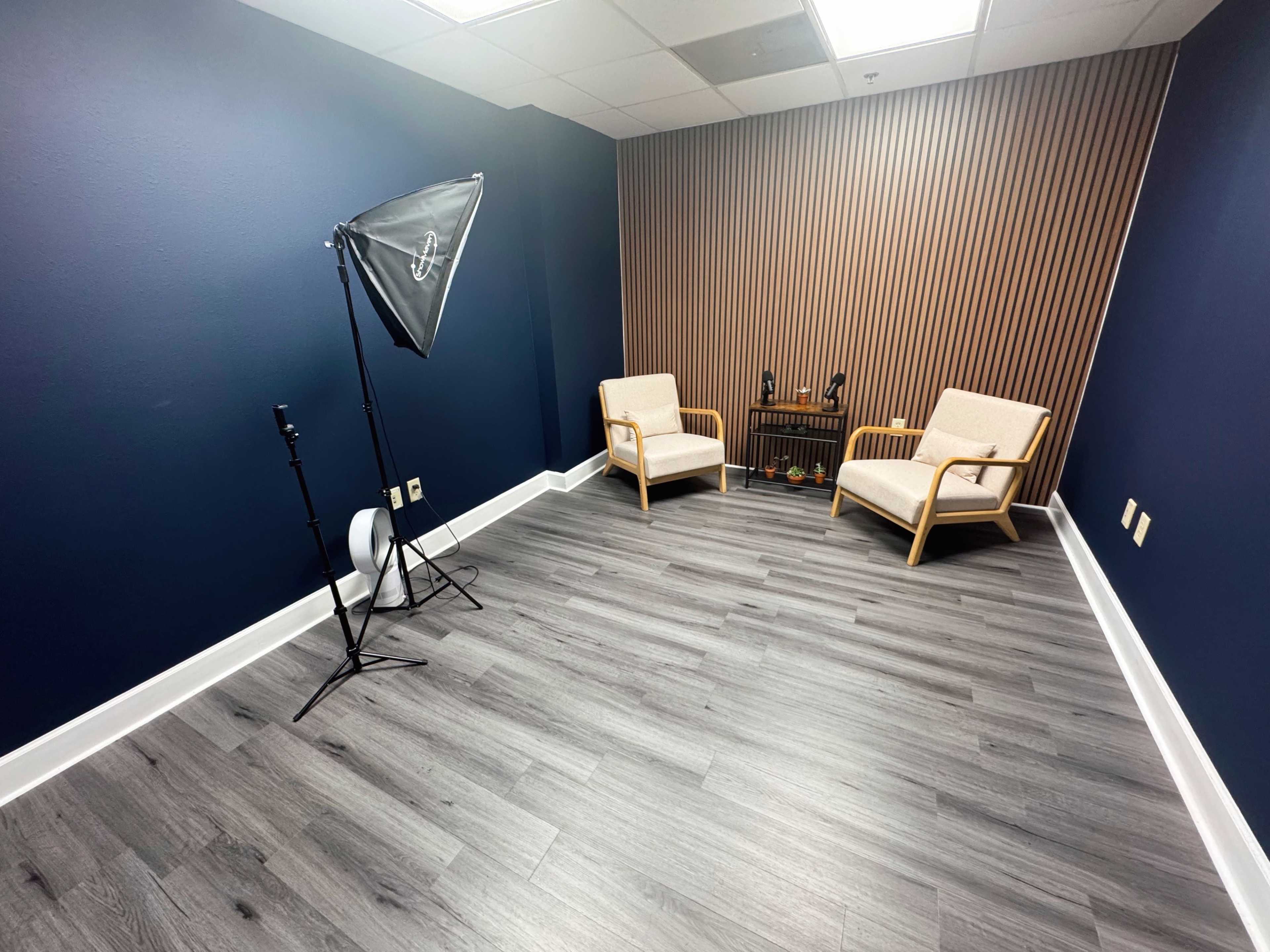The image shows a small room with two light-colored chairs, a dark wooden table, and a backdrop consisting of vertical wooden slats, accompanied by a lighting setup in one corner.