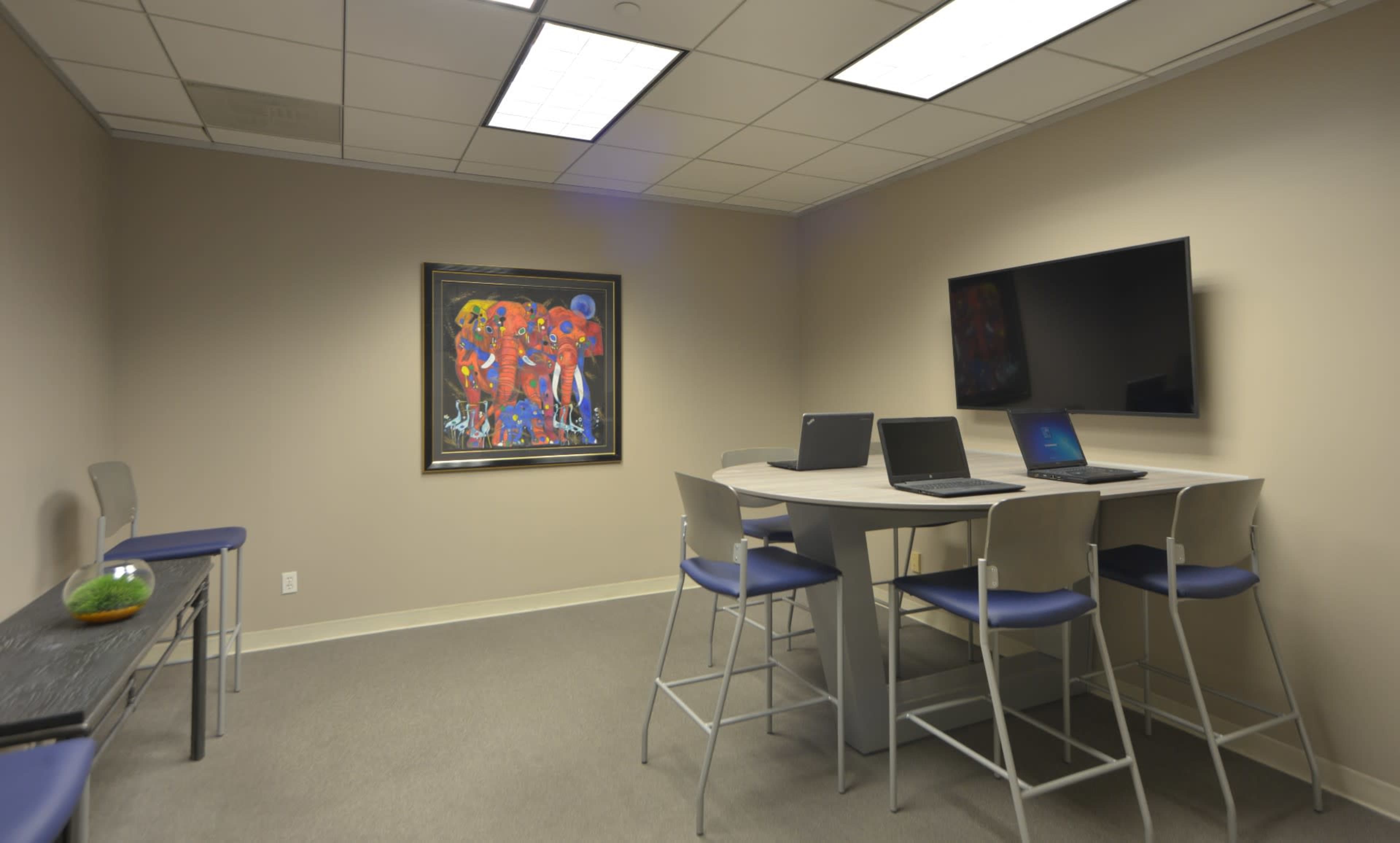A conference room with two high tables and chairs, featuring three laptops, a mounted television, and a vibrant piece of artwork on the wall.