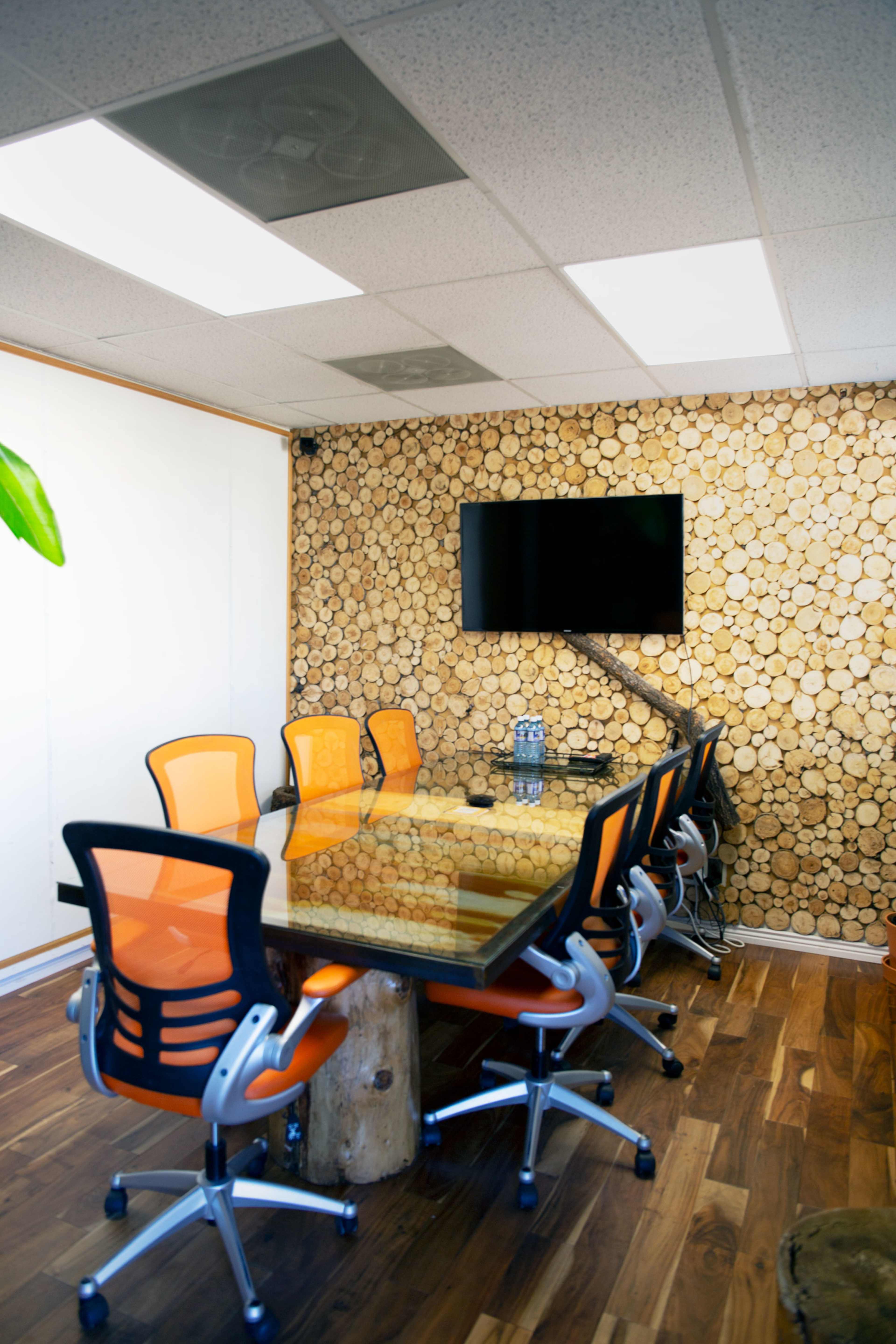 A conference room with a glass table surrounded by orange chairs, a wood-slice accent wall, and a television mounted on the wall.