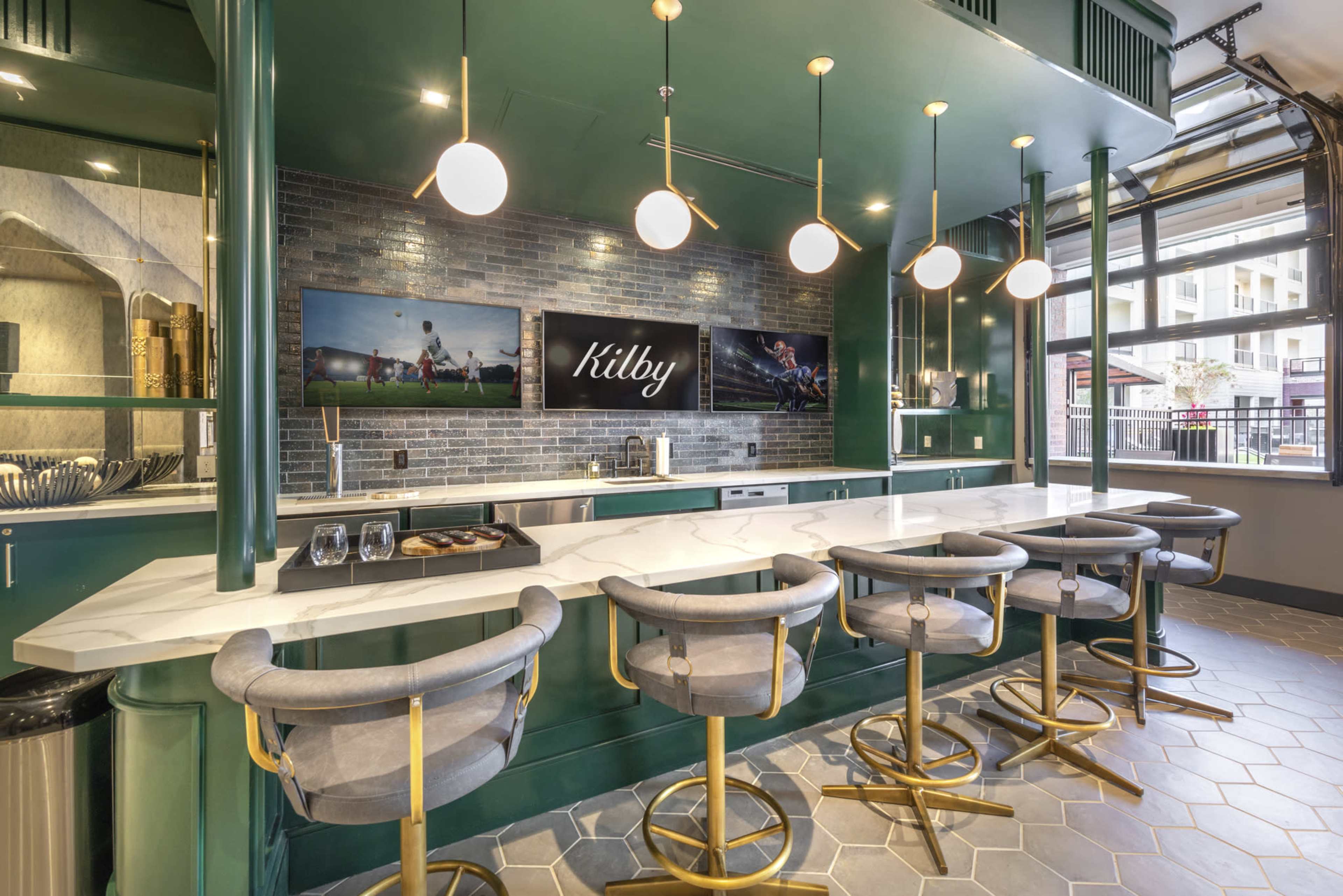 A modern kitchen bar area with a green accent wall, marble countertop, and gold-accented stools, featuring two large screens displaying the name "Kilby."