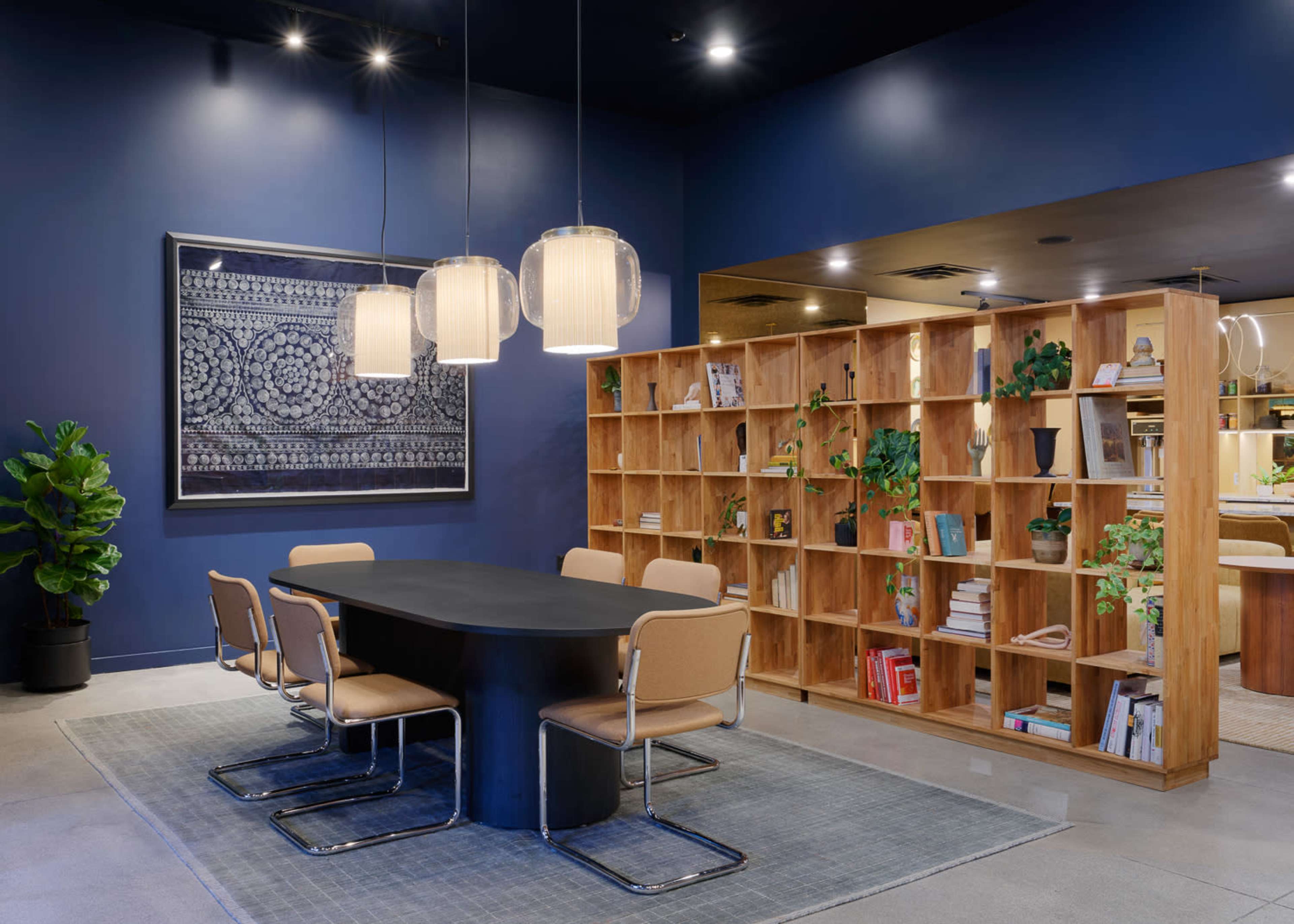 A modern conference room features a large oval table surrounded by beige chairs, with a wooden bookshelf filled with books and plants against a dark blue wall.
