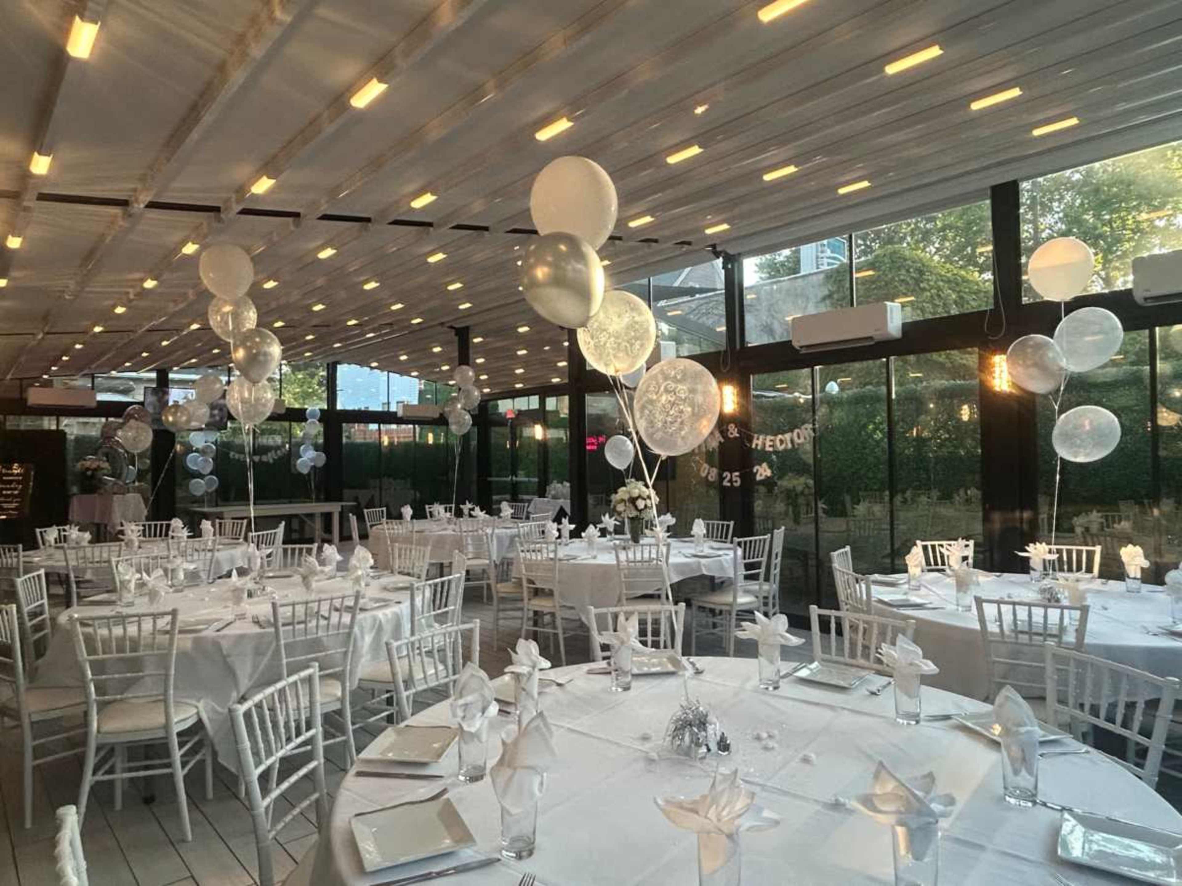 The image shows a beautifully arranged indoor dining space with round tables set with white tablecloths, elegant chairs, and decorative balloons suspended from the ceiling.