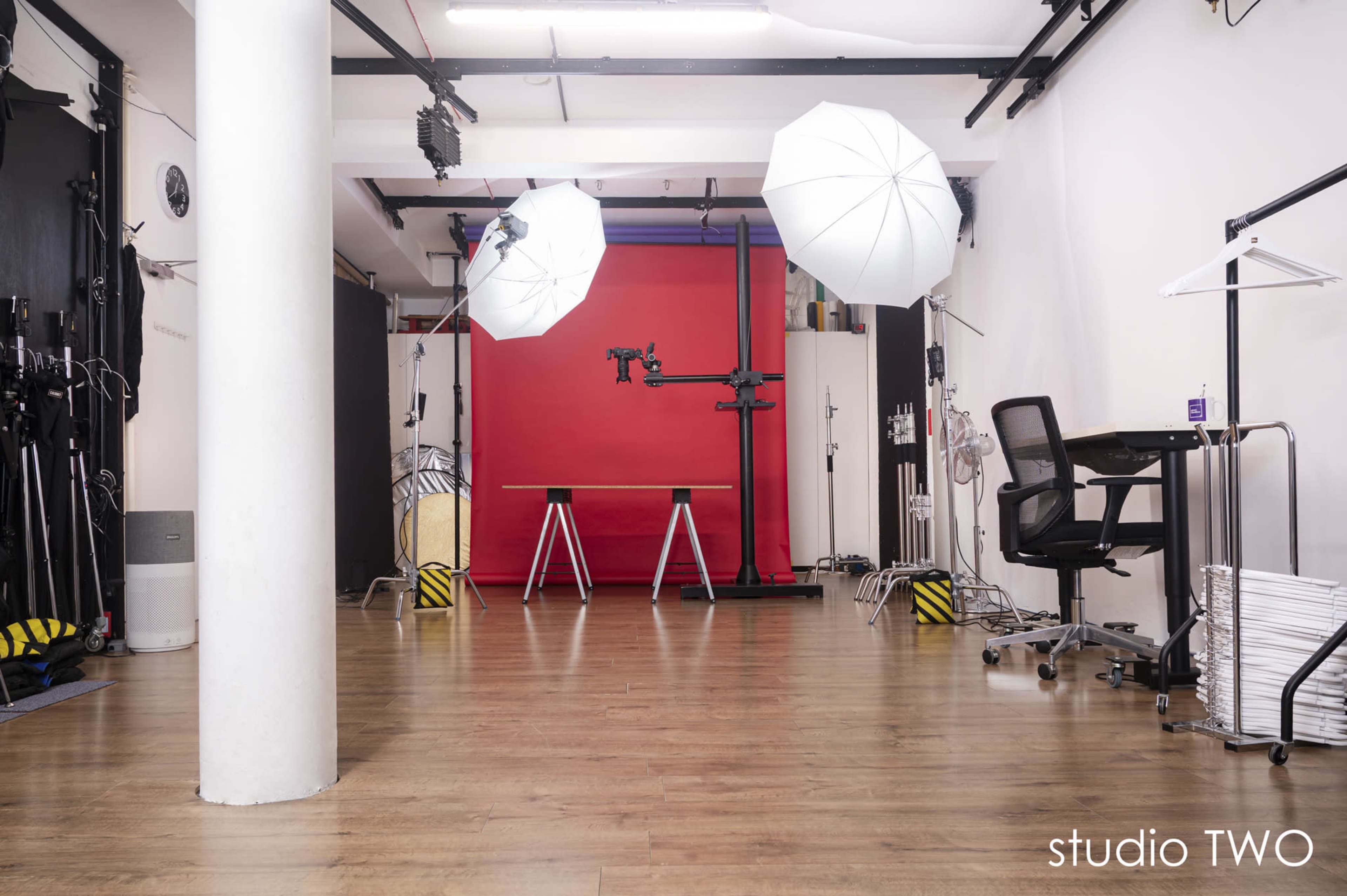 The image shows a photography studio with a red backdrop, two light stands featuring white umbrellas, and a chair and table arranged in the center.