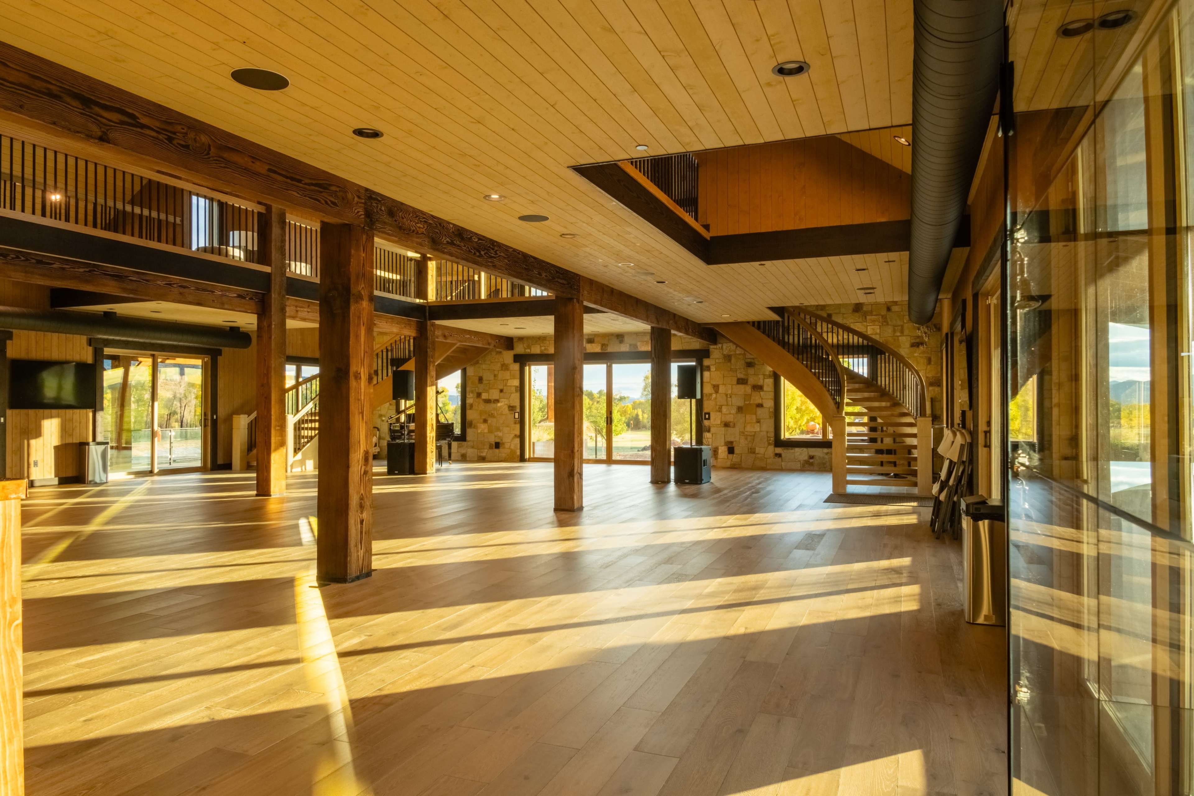The image shows a large, open interior space with wooden beams, a staircase, and abundant natural light streaming through large windows.