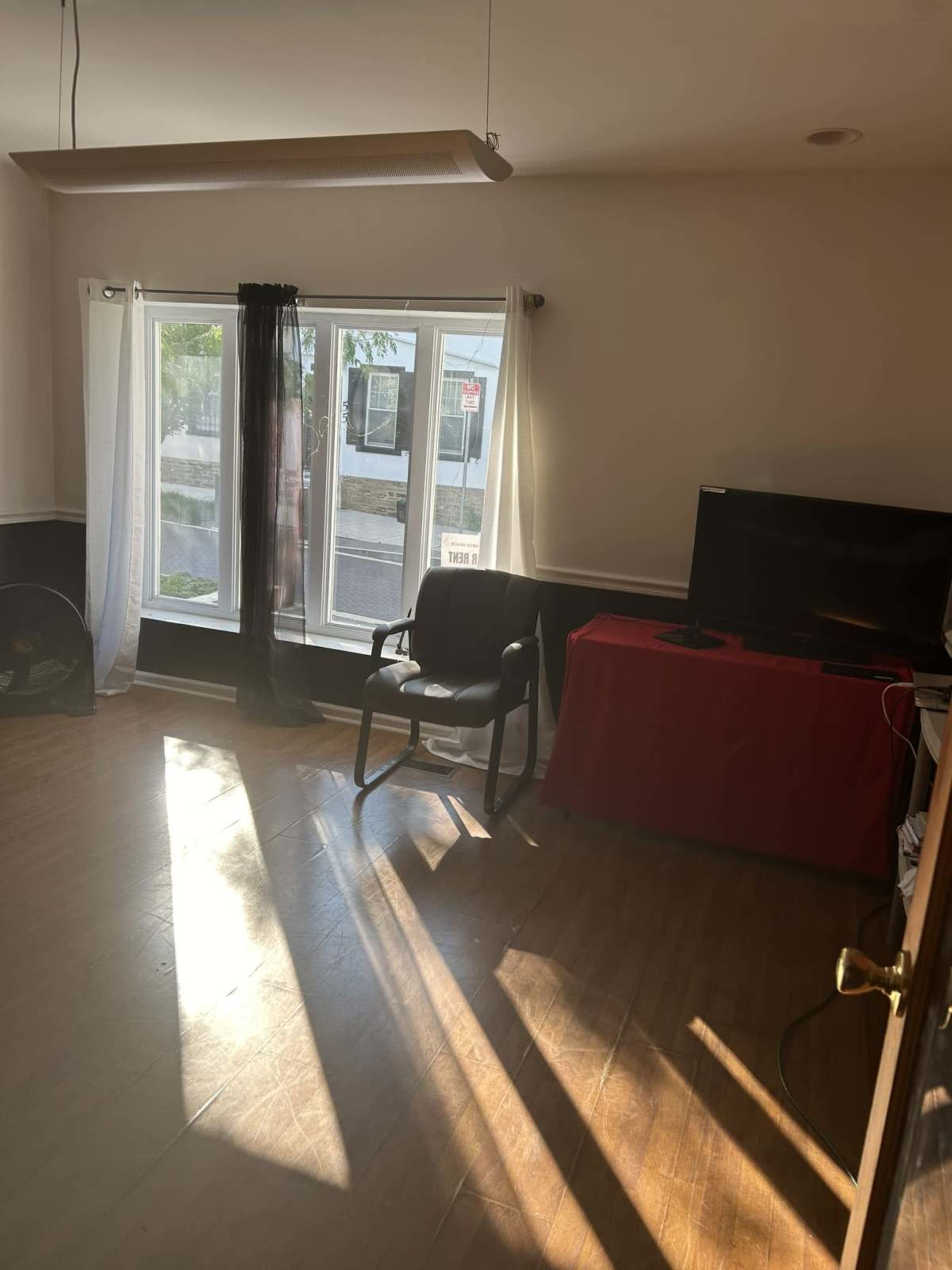 A room with wooden flooring features a window with sheer curtains, a black chair, a red tablecloth covering a table, and a television on a stand.
