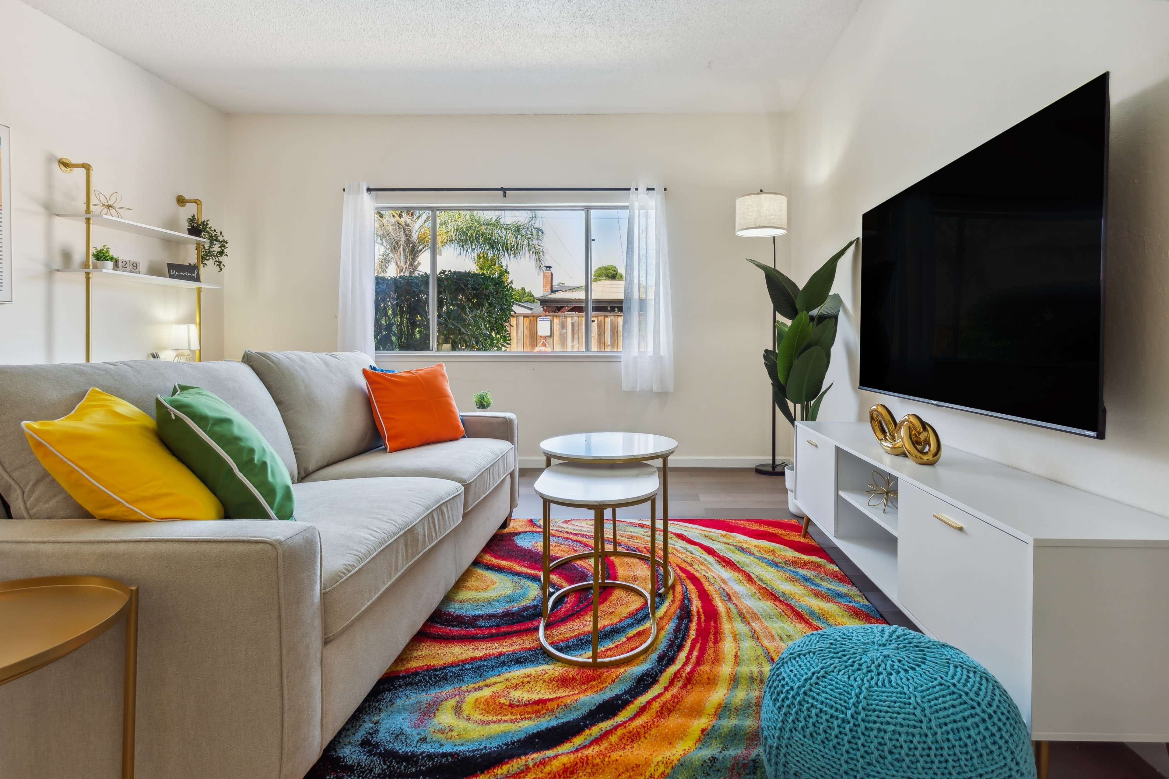 A bright living room features a beige sofa with colorful throw pillows, a round coffee table, a television on a white media console, and a vibrant, swirling area rug.