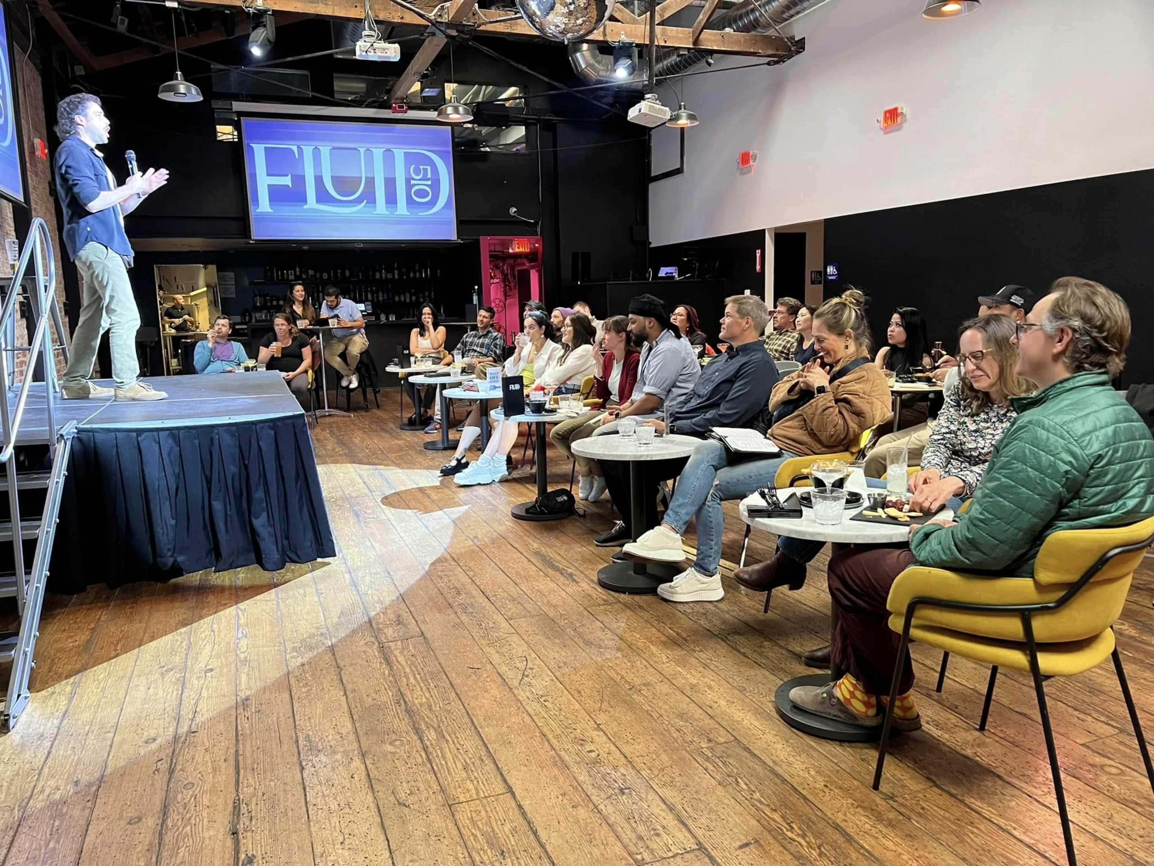 A speaker stands on a stage in front of an audience seated at tables in a modern event space.