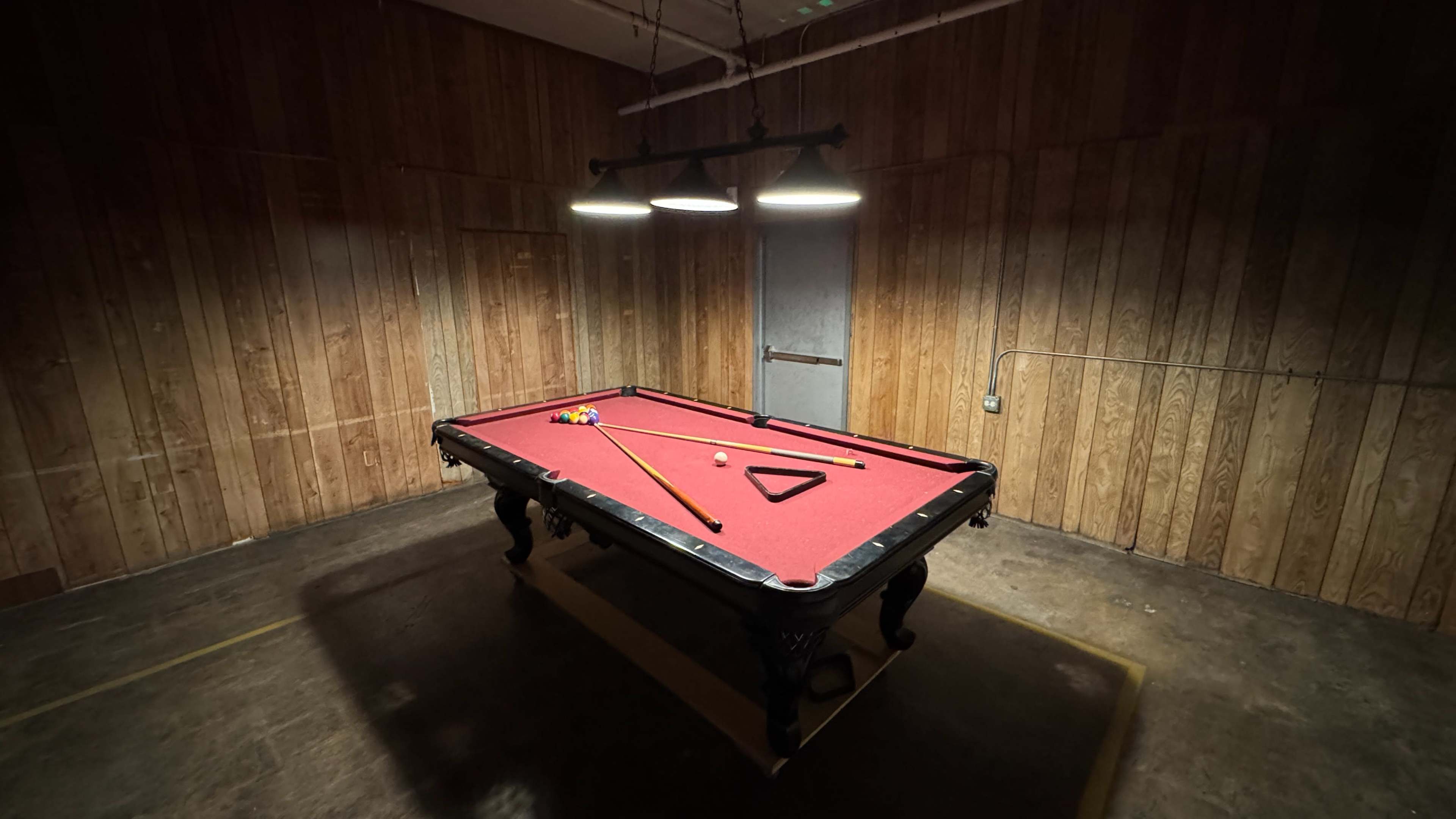 A red felt pool table is illuminated under three hanging lights in a dimly lit room with wooden paneling.