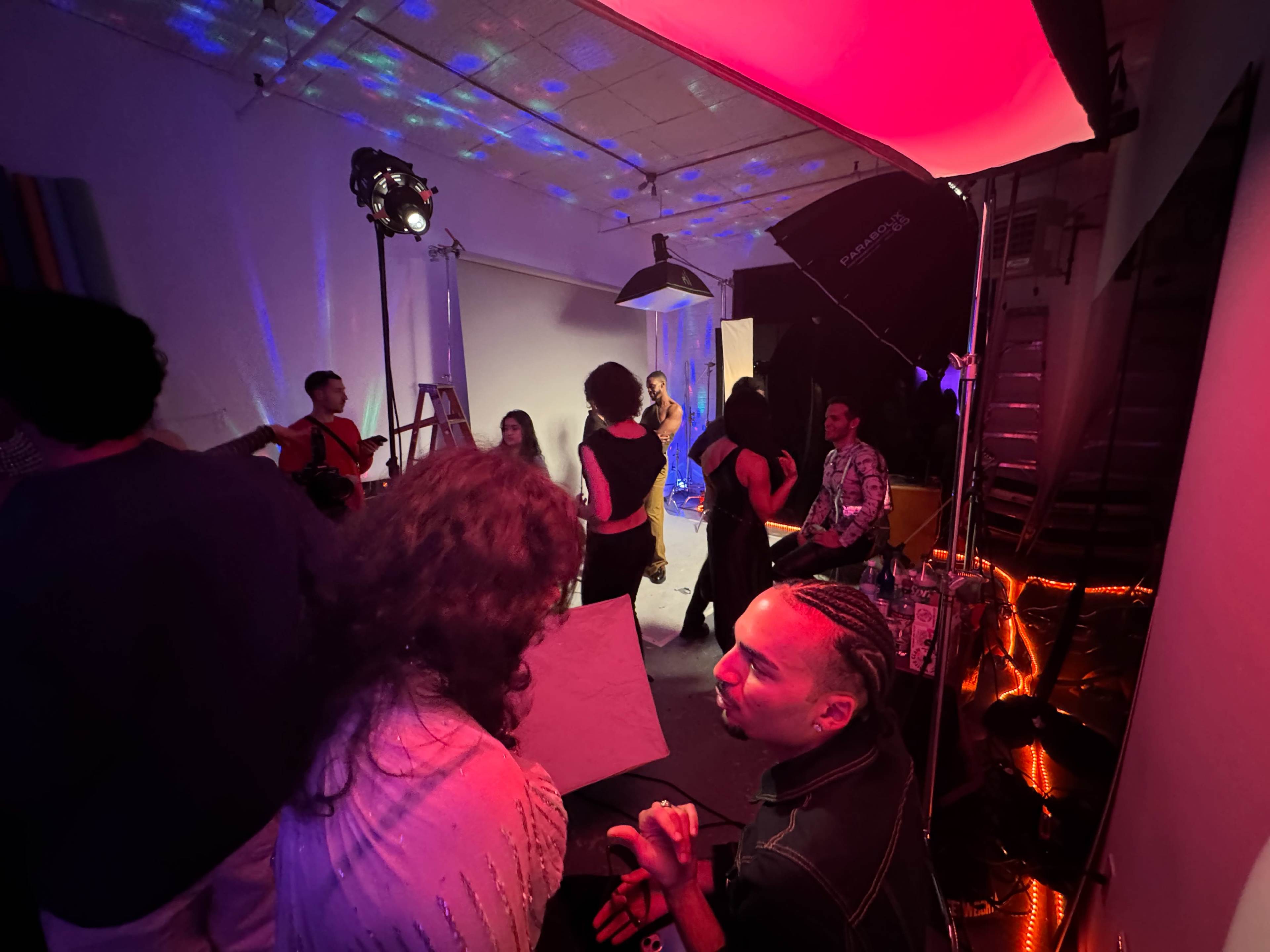 The image shows a vibrant studio setting with several people engaged in a creative photo shoot, illuminated by colorful lights and studio equipment.
